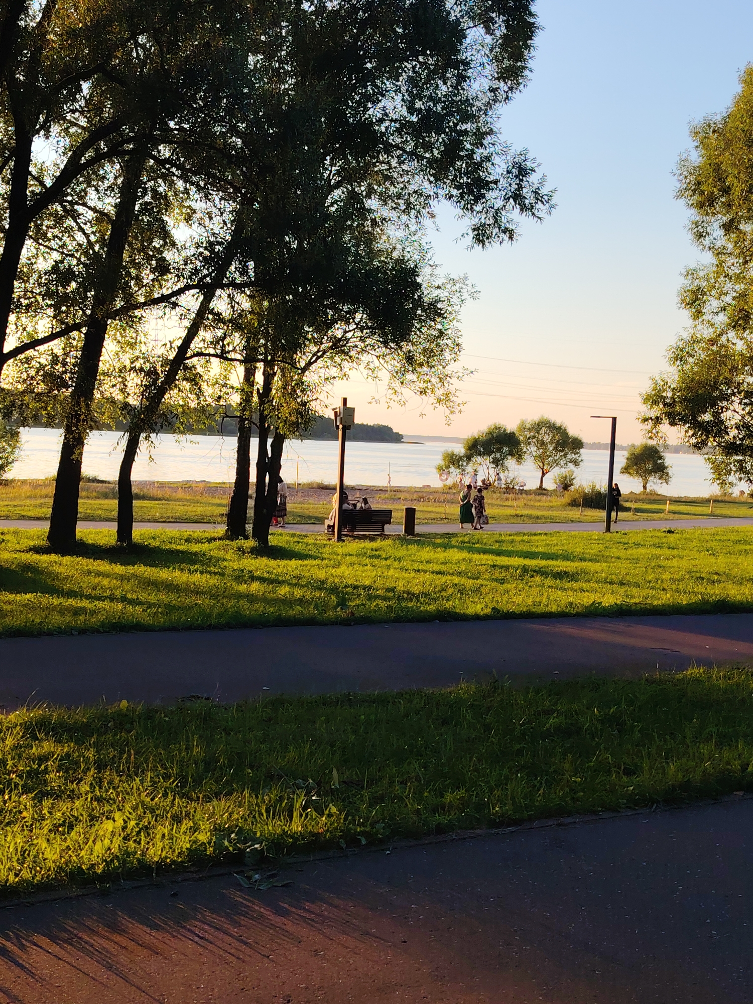 The image depicts a waterfront park area in Konakovo, Russia, bathed in the warm, low light of late afternoon or early evening. In the foreground, a paved pathway runs horizontally across the bottom of the frame, bordered by a strip of bright green grass. The grass is illuminated by the setting sun, casting long, distinct shadows across the pathway.
Further back, a wider paved walkway runs parallel to the water's edge. Along this walkway, several people are visible. Two individuals are seated on a dark bench, their backs to the camera. To their left, a group of three people are walking, one in a dark outfit and two in lighter, possibly patterned clothing. Further down the walkway, another person is walking away from the camera, silhouetted against the lighter background.
The middle ground features a lush expanse of green grass, interspersed with several mature trees that cast dappled shadows. Beyond the grassy area and walkway lies a body of water, likely the Ivankovo Reservoir, its surface reflecting the soft, pale sky. The distant shoreline is dotted with trees, and the sky is a clear, pale blue, transitioning to a lighter hue near the horizon. A lamppost stands on the right side of the walkway. The overall impression is one of a peaceful, recreational public space.