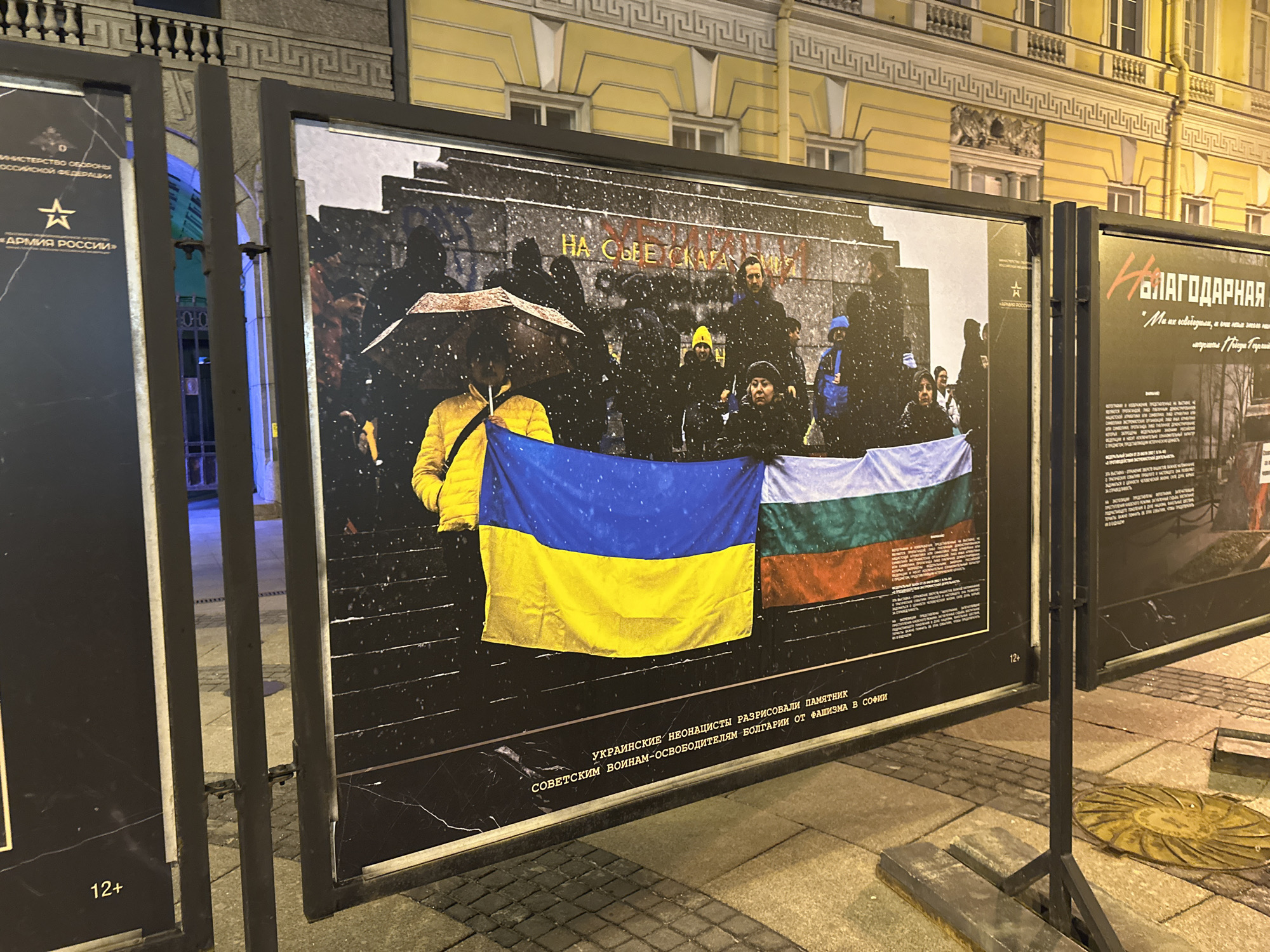 The image shows an outdoor display of photographs in Saint Petersburg, Russia. The photograph on display depicts a group of people holding the flags of Ukraine and Bulgaria in front of a monument. The text accompanying the photograph states that Ukrainian neo-Nazis vandalized the monument to Soviet soldiers-liberators of Bulgaria from fascism in Sofia. The background includes a snow-covered cityscape. The display is set on a paved surface, and the surrounding environment includes a building and street lighting.