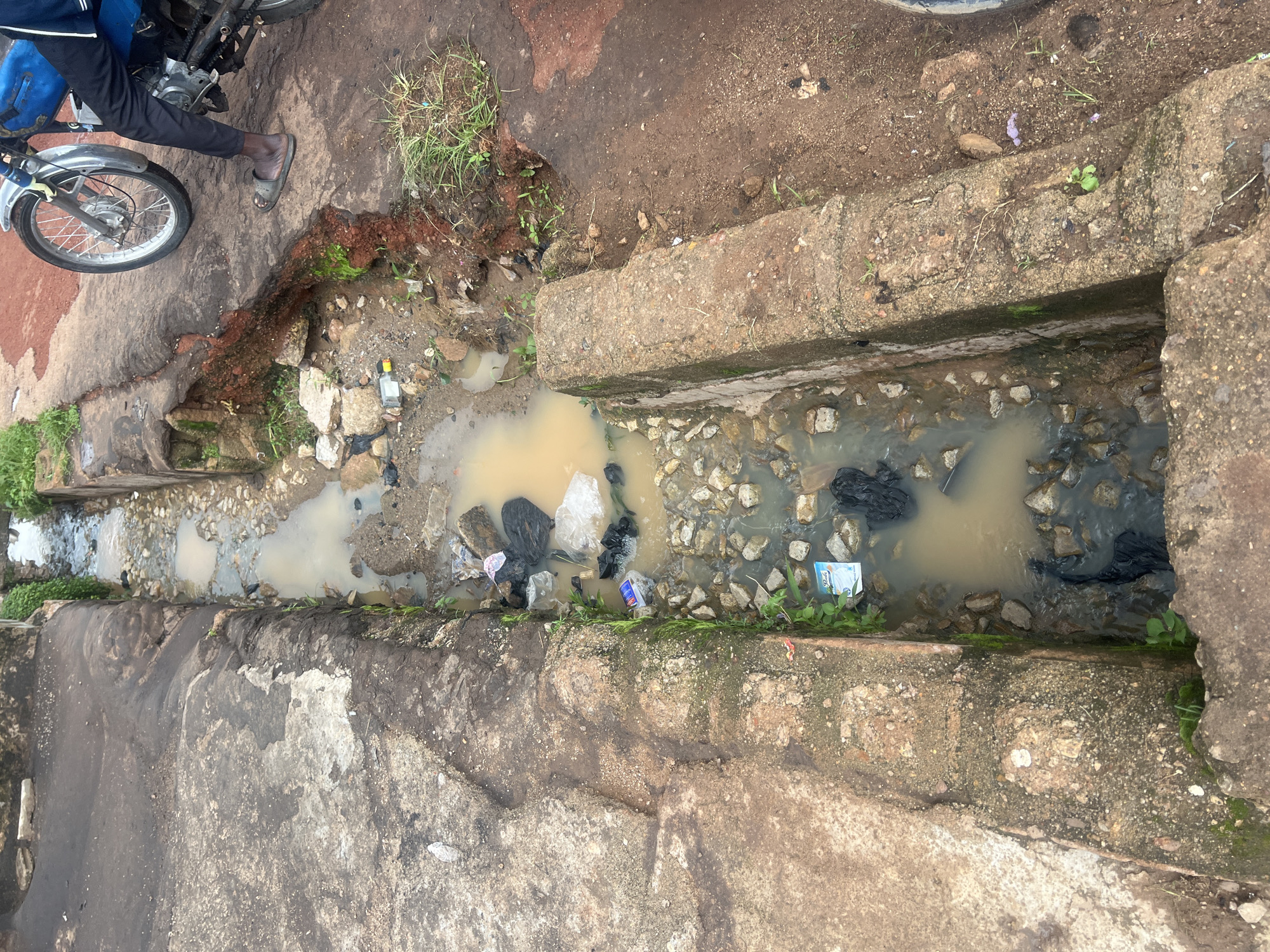 The image shows a dirty, shallow drainage channel filled with murky water and debris. The channel is lined with concrete blocks and appears to be alongside a dirt or gravel road. Various pieces of trash, including plastic bags and bottles, are visible in and around the water. Some green vegetation is growing in and around the edges of the channel.
In the upper left portion of the image, a person's legs and feet are visible. They are wearing dark pants and a gray sandal. The person is standing next to a motorcycle, with the front wheel and part of the engine visible.
The scene suggests an outdoor environment, likely in a somewhat neglected urban or semi-urban area. The lighting appears to be natural daylight, and there are no strong indicators of specific weather conditions beyond the presence of water in the drainage. The overall impression is one of disrepair and poor sanitation. There is a visible piece of packaging with text that appears to read "PUNCH" in blue letters on a white background.