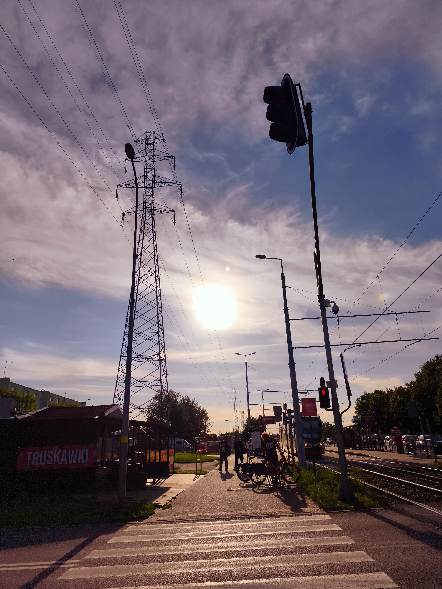 This image captures a street scene in Gdańsk, Poland, specifically at a tram stop near a high-voltage power line pylon. The sun is low in the sky, casting long shadows and creating a warm, golden light, suggesting either sunrise or sunset.
In the foreground, a pedestrian crossing with white stripes is visible on an asphalt road. To the left of the crossing, a small, dark-colored kiosk or shed with a red banner displaying the word "TRUSKAWKI" (Polish for "strawberries") is present. Behind the kiosk, grass and some trees are visible.
Further into the scene, a paved pedestrian walkway runs alongside tram tracks. Several people are standing on this walkway, some with bicycles. Two bicycles are leaning against each other. In the background, a tram can be seen approaching or stopped at the station. A traffic light, showing red, is mounted on a pole. Another pole supports a CCTV camera. High-voltage power lines crisscross the sky, with a large, lattice-style pylon dominating the left side of the frame. The sky is filled with scattered clouds, tinged with purple and pink hues from the low sun. Buildings and trees form the backdrop on the far left and right sides of the image. The overall impression is an urban environment during a colorful twilight.