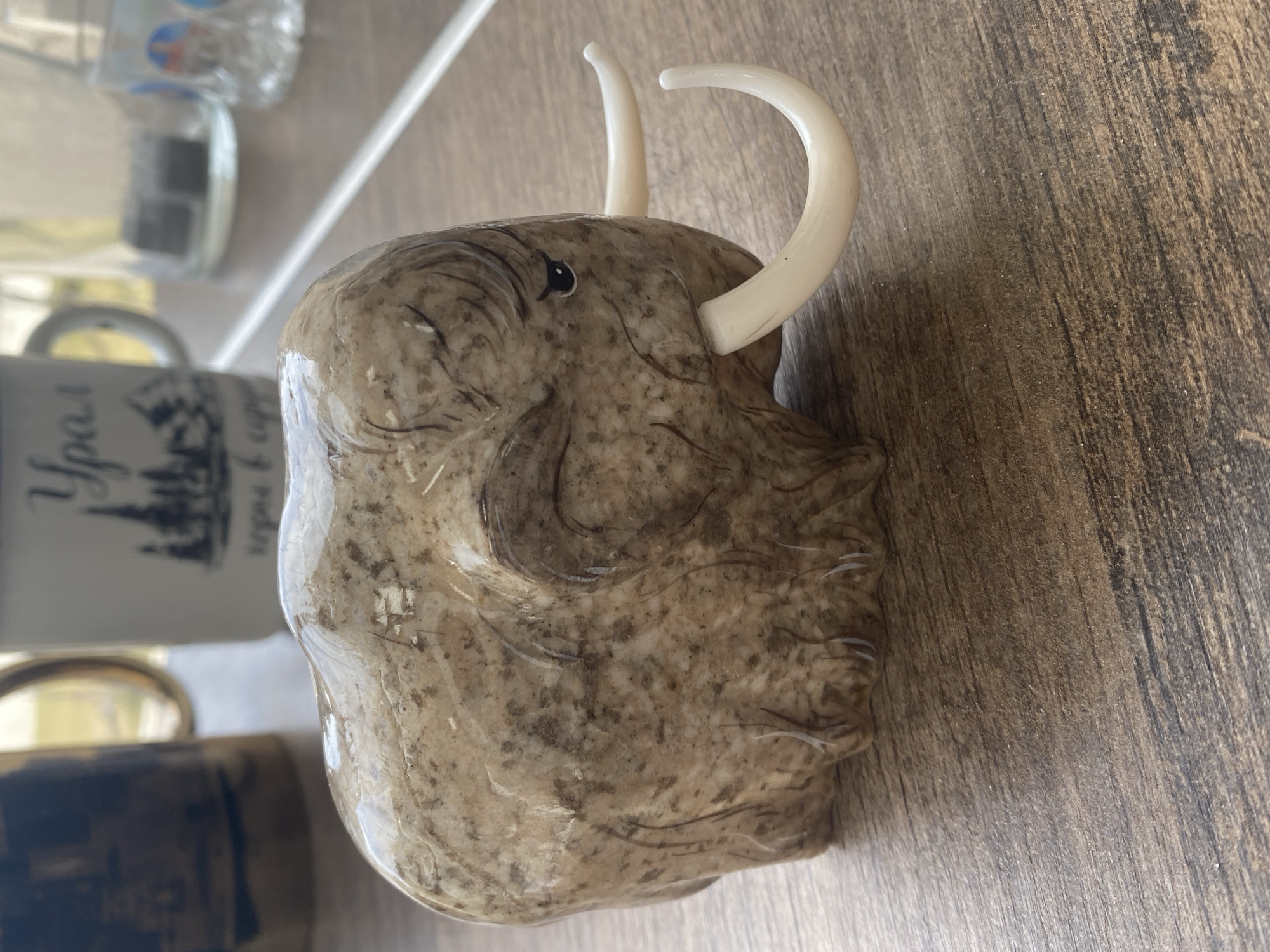 A ceramic mug shaped like a mammoth is on a wooden table. The mug is predominantly brown with lighter, speckled detailing to simulate fur. It has a small, dark eye and two white tusks that curl upwards. To the left of the mammoth mug, slightly out of focus, is a mug that says "Урал" on it. Behind the mugs, there is a partially visible clear plastic bottle. The surface the items rest on is a brown wooden surface. It is unknown if this is in Yekaterinburg, Russia.