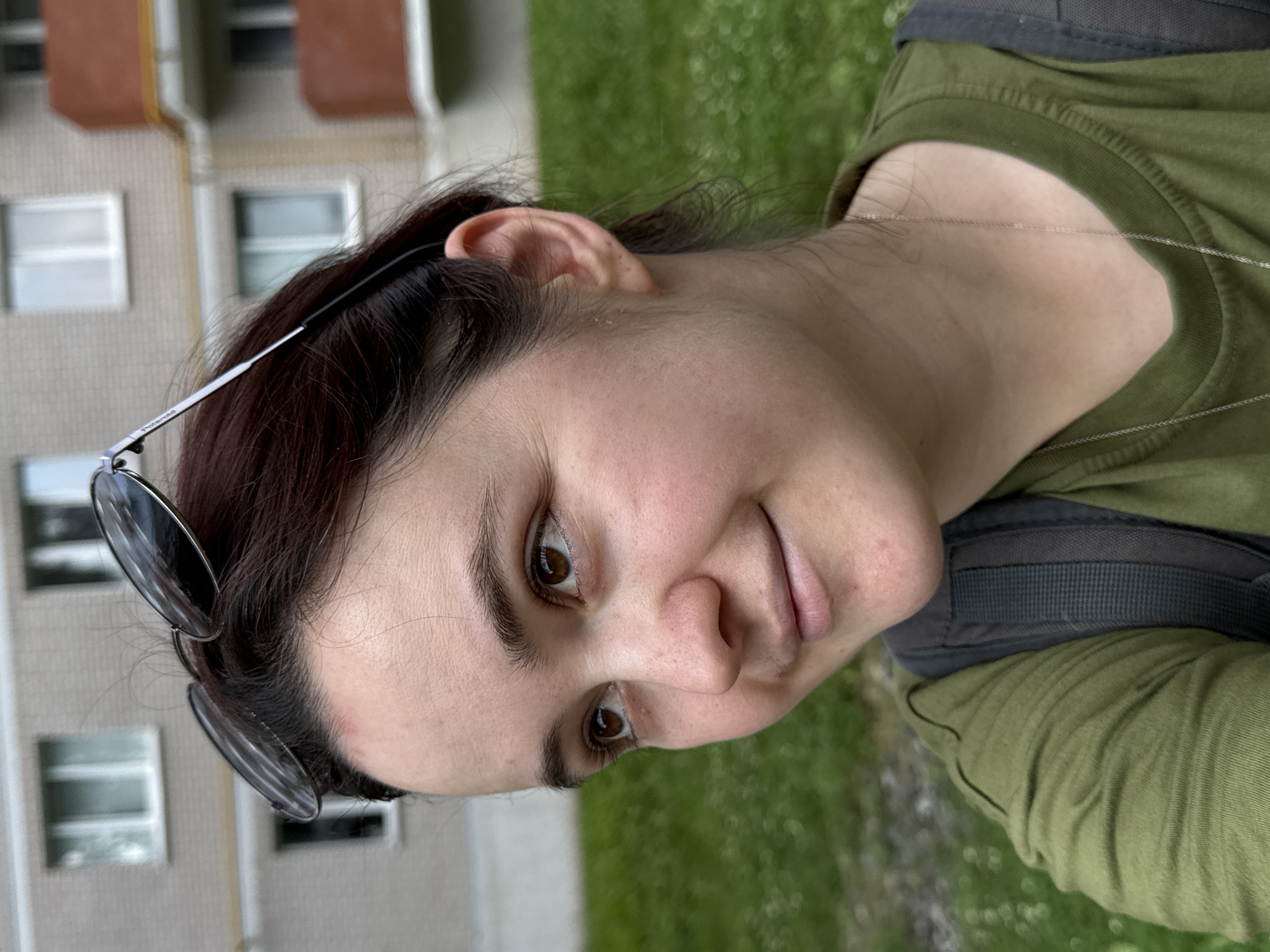 The image is a close-up, medium shot of a young Caucasian woman with brown hair and brown eyes, looking to her left. She is wearing a green t-shirt and a backpack, with sunglasses perched on her head. A slight red mark is visible on her forehead, and some acne is present on her chin. The background suggests an urban environment, with a brick apartment building visible behind her. The ground is covered in grass, indicating an outdoor setting. This scene is likely located in the city of Izhevsk, Russia, given the architectural style of the building and the general environment.