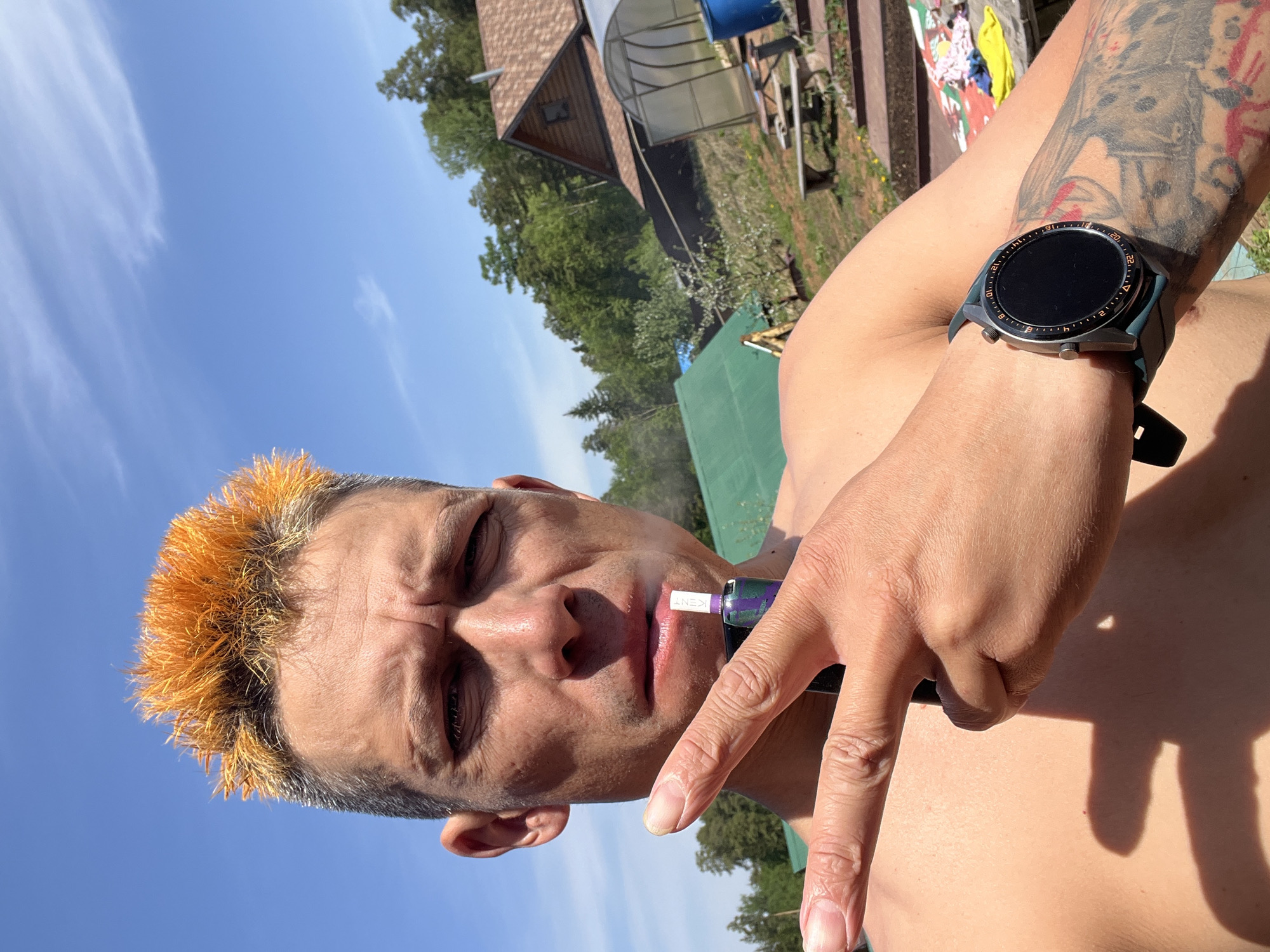 A man with a partially shaved head and orange-dyed hair is holding an electronic cigarette to his lips. He is wearing a smartwatch on his left wrist, which is tattooed. The background shows a blue sky, trees, a wooden house, and a greenhouse. The photograph was taken during the day. I cannot ascertain the location from the image.