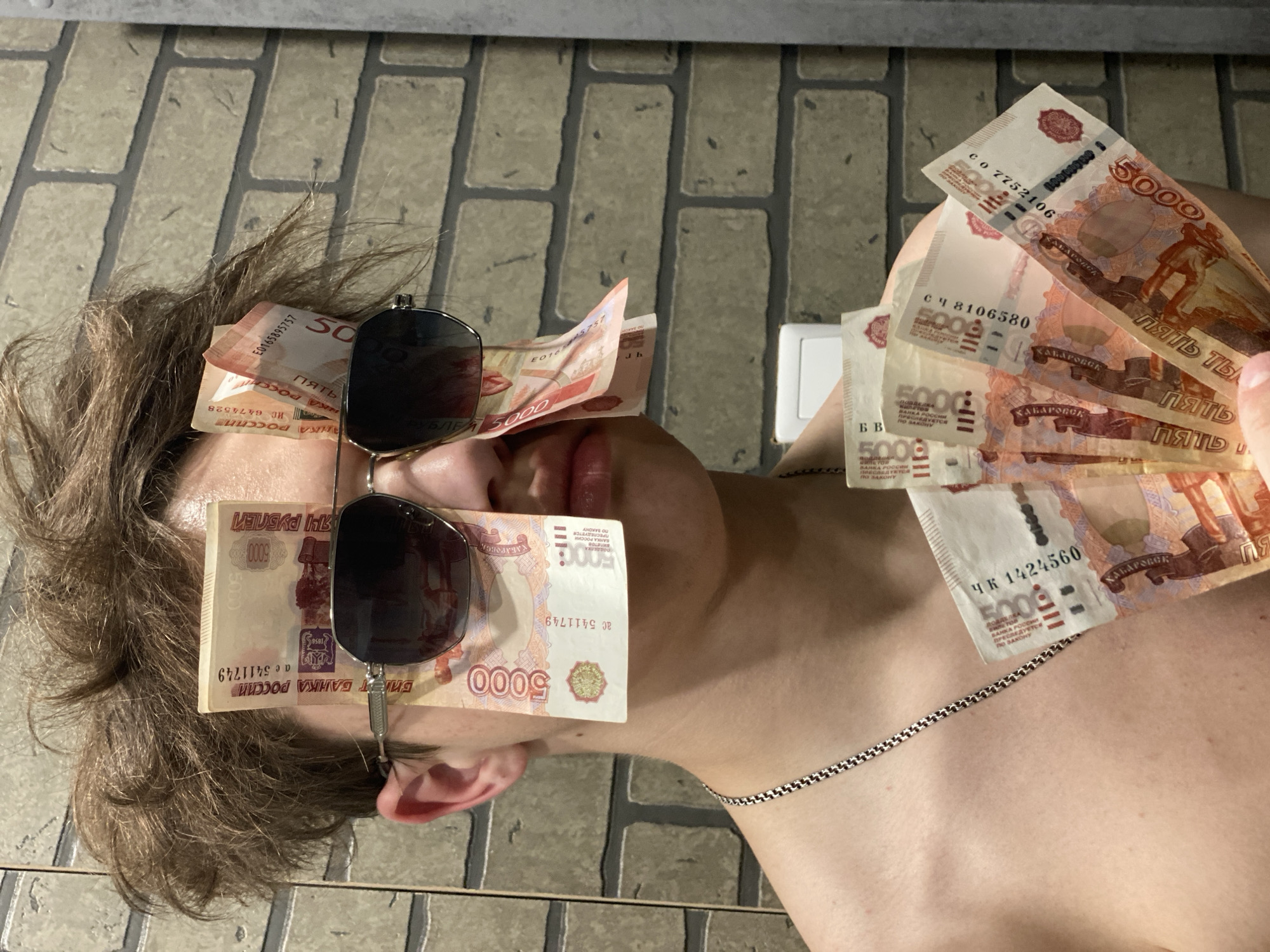 A person is depicted lying on their back on a light brown tiled surface, possibly an indoor floor or an outdoor patio, under daylight conditions. The individual's face is largely obscured by a pair of sunglasses and several 5000 Russian Ruble banknotes placed directly over their eyes. More banknotes are fanned out and held on their chest and shoulder area.
The subject has brown hair and is wearing a silver-colored chain necklace. Their lips are visible, slightly parted. The banknotes are predominantly orange and red, featuring the value "5000" and the text "ПЯТЬ ТЫСЯЧ РУБЛЕЙ" (Five Thousand Rubles), "БИЛЕТ БАНКА РОССИИ" (Bank of Russia Note), and "ХАБАРОВСК" (Khabarovsk), indicating the city depicted on the currency. Phrases like "ПРЕСЛЕДУЕТСЯ ПО ЗАКОНУ" (Punishable by Law) are also visible, referring to counterfeiting. Serial numbers such as E0165895757, ИС 6474528, С0 7752106, СЧ 8106580, ЧК 1424560, and АС 5411749 are legible on the various bills. The scene, set in Sochi, Russia, suggests a display of wealth or a casual interaction with a significant amount of currency.