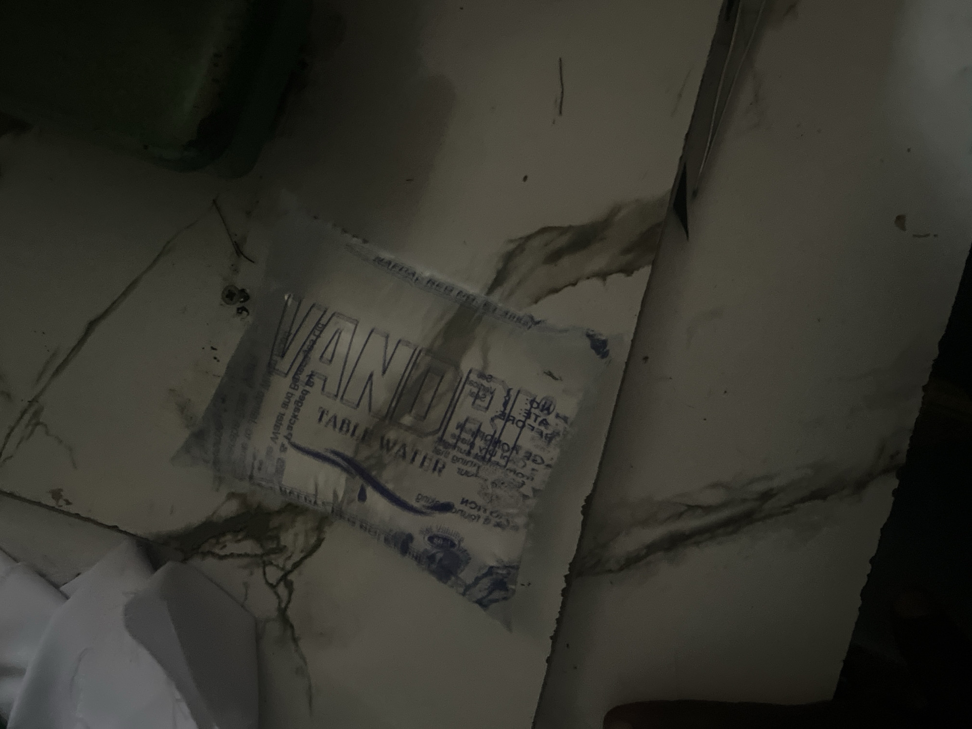 The image shows a small packet of table water, likely a single serving. The packet is clear plastic with blue text indicating "TABLE WATER" and other branding. It is lying on a white, marbled surface that appears to be a countertop or table. The lighting is dim, with shadows casting across the surface. There are some dark, organic-looking stains on the surface near the packet. In the background, a green object, possibly a bottle or container, is visible in shadow. A small piece of white paper or plastic is also in the upper left corner of the image. The overall scene appears to be an indoor setting in Jalingo, Nigeria, possibly a domestic or commercial environment.