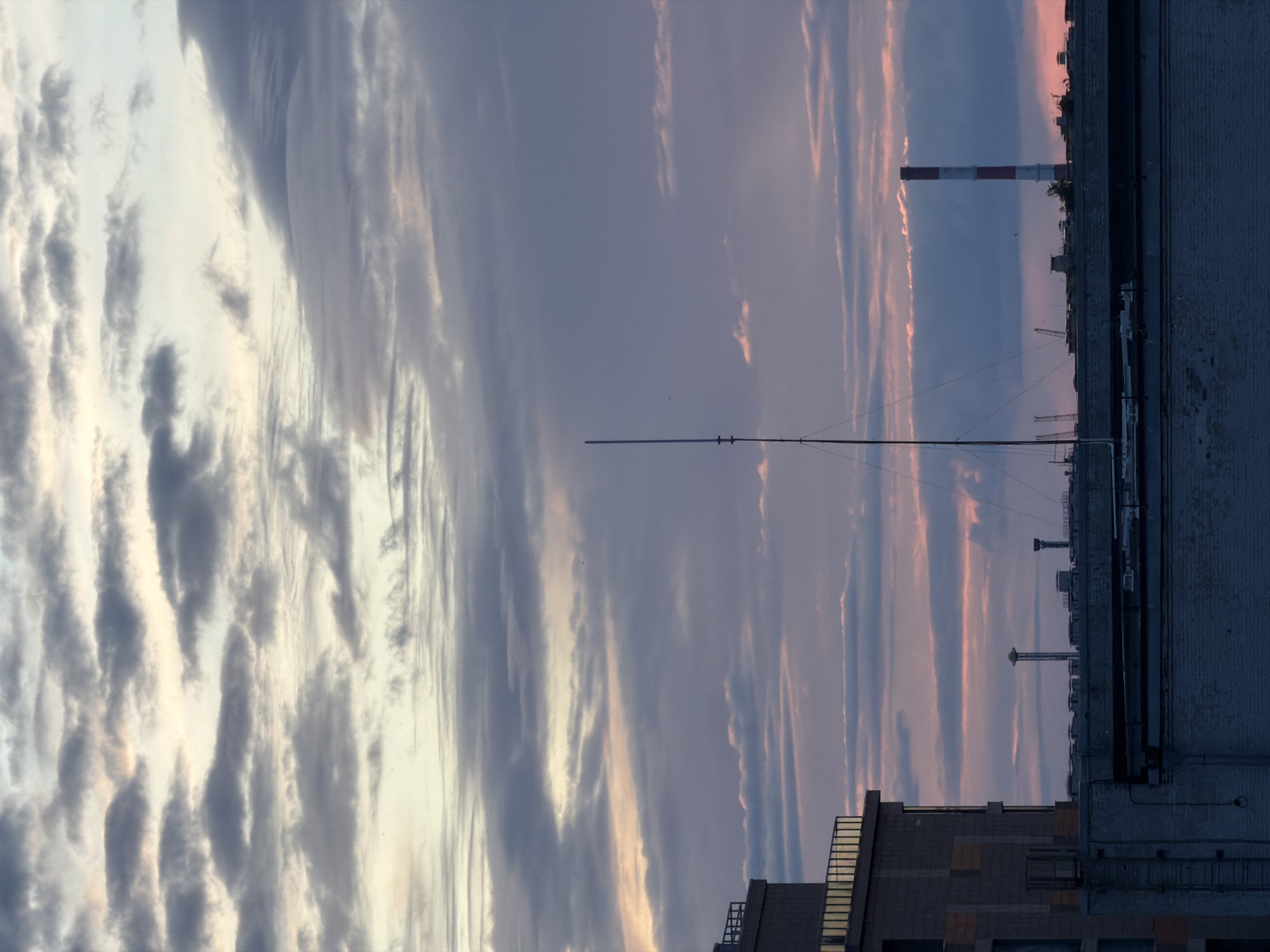 This is a vertical, medium shot of a cloudy sky above a cityscape in an unknown location in Russia. The sky is filled with dramatic, layered clouds in shades of gray, white, and muted pink, suggesting either sunrise or sunset. A tall, slender antenna stands prominently in the center of the frame, reaching into the clouds. In the lower portion of the image, the rooftops of several buildings are visible, including a modern building with railings on the left and a brick building with what appears to be an industrial chimney with red and white stripes on the right. Other structures and a hint of greenery are also present on the rooftops. The overall atmosphere is somewhat industrial and urban, with the natural beauty of the sky providing a striking contrast. There are no people or animals visible in the image.