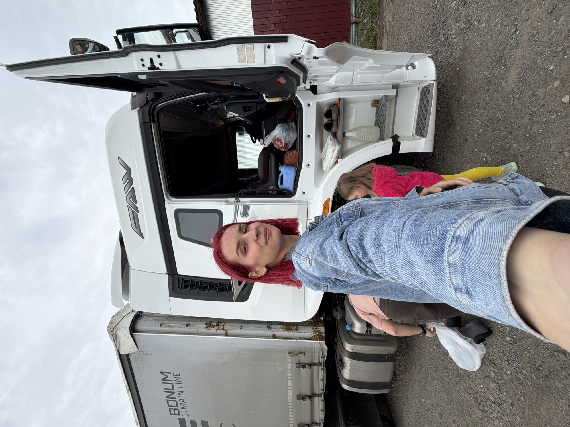 This portrait-oriented photograph captures a woman and a child beside a large white semi-truck and trailer under an overcast sky. The scene is set outdoors on a gravel surface, with a reddish-brown metal-clad building visible in the background.
In the foreground, a woman with short, bright red hair, wearing a denim jacket over a light-colored top, poses for the camera with a subtle smile. She appears to be holding the camera for a selfie or taking the photo from a low angle. Her left arm is visible in the bottom right corner of the frame.
To her right, partially obscured by the truck and her body, a child in a pink long-sleeved top and bright yellow leggings is seen crouching or kneeling near the truck's side. The child's upper body, legs, and feet in light-colored sneakers are visible, and a white plastic bag rests on the ground nearby.
The central subject is a white FAW brand semi-truck. Its driver's side door is open, revealing the cab's interior, which contains various items including bags and a blue container. External storage compartments near the front wheel are also open, housing items like shoes, a white plastic bottle, and what appears to be another plastic bag. The truck is connected to a silver-colored trailer with "BONUM MAIN LINE" written on its side. The trailer's upper frame shows visible rust.
The image suggests a stop or break during a journey, possibly at a truck stop or yard in Kamyennoye, Russia. The overcast sky indicates daytime without direct sun.