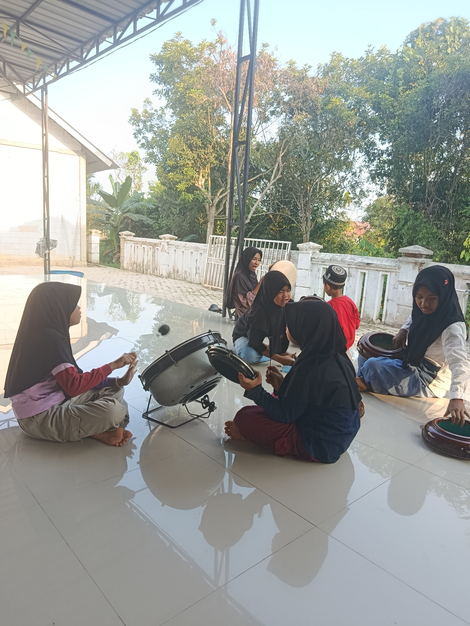 In Swarga Bara, Indonesia, a group of children, most of whom are wearing hijabs, are gathered on a tiled outdoor floor beneath a covered patio. In the foreground, to the left, a young girl in a pink shirt and grey pants sits cross-legged, playing a large drum with drumsticks. To her right, another girl in a black hijab and dark blue pants also appears to be playing a drum, holding a smaller drum in her lap. Further to the right, a young boy in a red shirt and a patterned kufi is visible, and beside him, a girl in a black hijab and blue jeans is playing a drum. Another girl in a white shirt and black hijab is seated in the far right of the frame, also seemingly engaged in playing a drum. Several drums are present in the scene, with some children holding them and others playing them. The background shows a white building, a white railing, and lush green trees. The lighting suggests it is daytime, possibly late afternoon, with shadows cast on the floor.