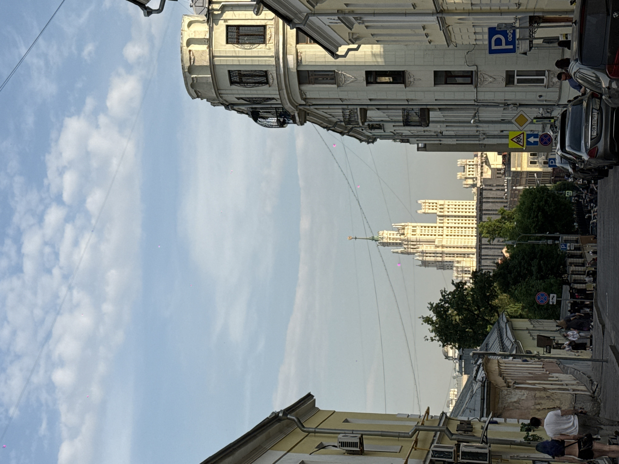 The image depicts a street view in Moscow, Russia, facing the Kotelnicheskaya Embankment Building, a Stalinist skyscraper. The buildings on either side of the street are of varying styles and heights. On the right side of the street, cars are parked, and there are people walking on the sidewalk. A street sign is visible. On the left, a cream-colored building with rounded corners is prominent. The sky is light blue with clouds. Power lines are visible above the street.