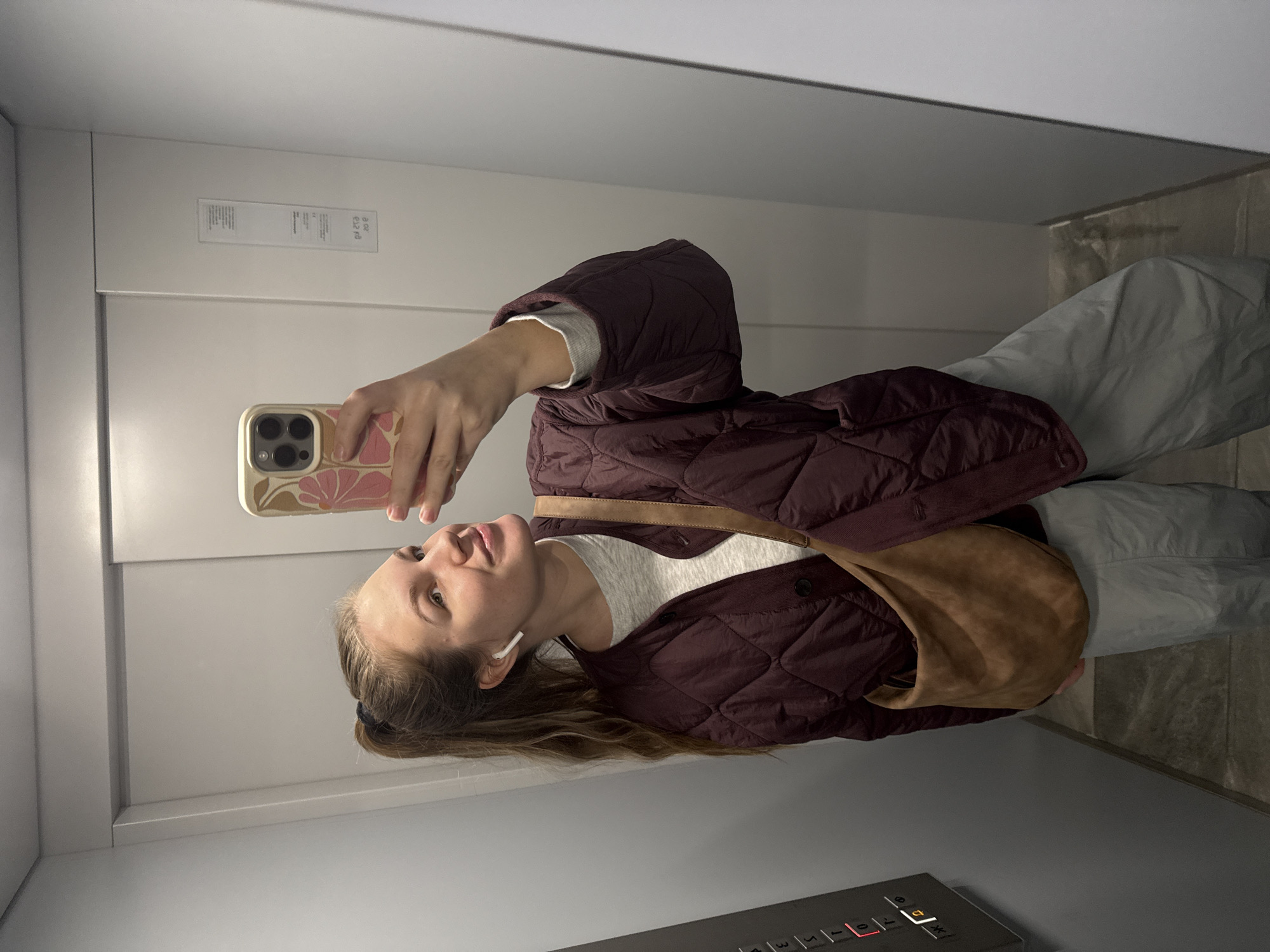 A woman stands in an elevator, taking a selfie with her smartphone. She wears a maroon quilted jacket, a light gray sweater, and light gray pants, with a brown leather shoulder bag. She has an earpiece in her left ear. The elevator interior is predominantly gray with a control panel visible at the bottom. The elevator door has small rectangular plates with writing on it. The location is likely in a building within the urban municipality of Vozdovac in Serbia.