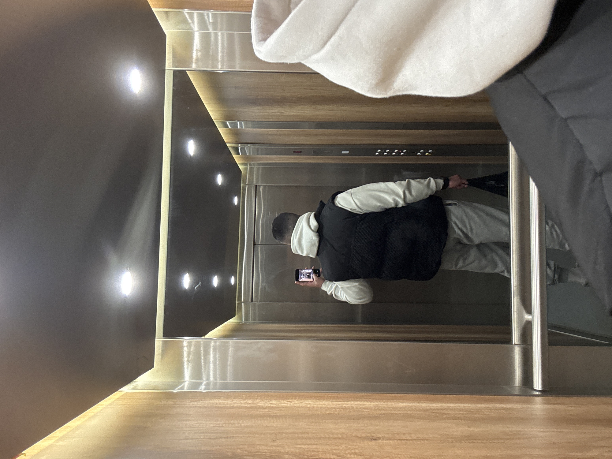 A person is taking a photo of themselves in a mirrored elevator. The elevator has a black reflective wall with a row of lights. The buttons are located on a wood-paneled wall. The person in the reflection is wearing a gray hooded sweatshirt and a black vest. Another person is partially visible on the right side of the image, wearing a white hoodie and black jacket. The location appears to be an indoor space in Sofia, Bulgaria, but the details of the city are not visible.