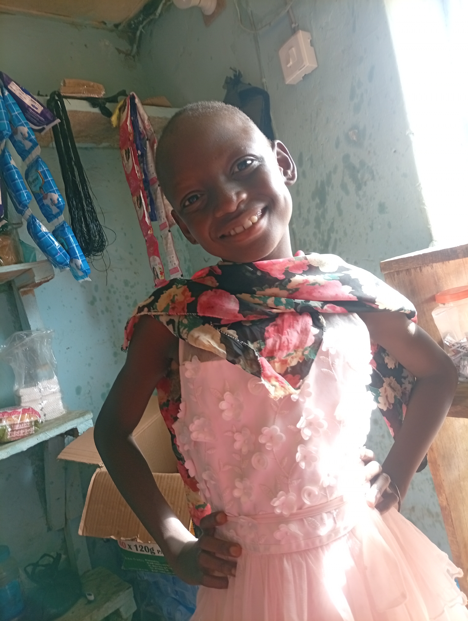A young person, possibly a child, with a shaved head is wearing a pink dress with floral appliqués and a floral shawl draped over their shoulders. They are smiling at the camera with hands on their hips. The background includes a cluttered room with items such as wrapped goods hanging from a shelf, a cardboard box, and personal items. The location is in Unknown, Unknown.