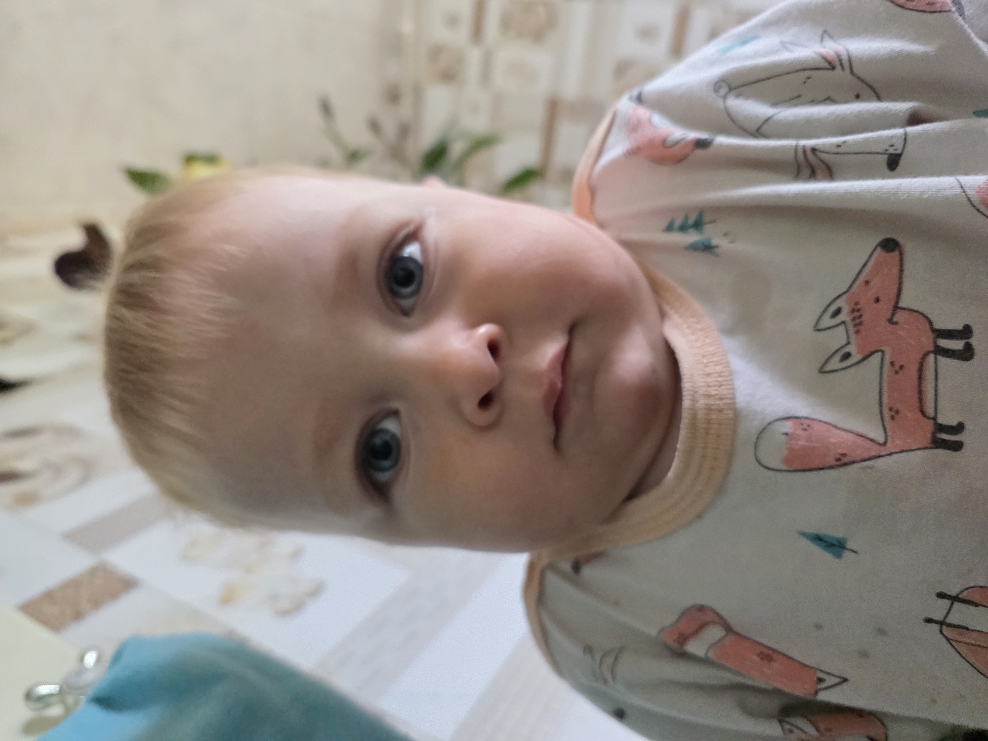 A close-up, slightly rotated image features a baby with fair skin, blonde hair, and blue eyes. The baby is wearing a light-colored onesie patterned with coral-colored foxes and blue trees. The baby's face is the primary focus, with the nose and mouth in the center. The background is out of focus, but hints of a tiled floor, foliage, and a wall can be seen. The photo was taken in the city of Chaikovsky, Russia.