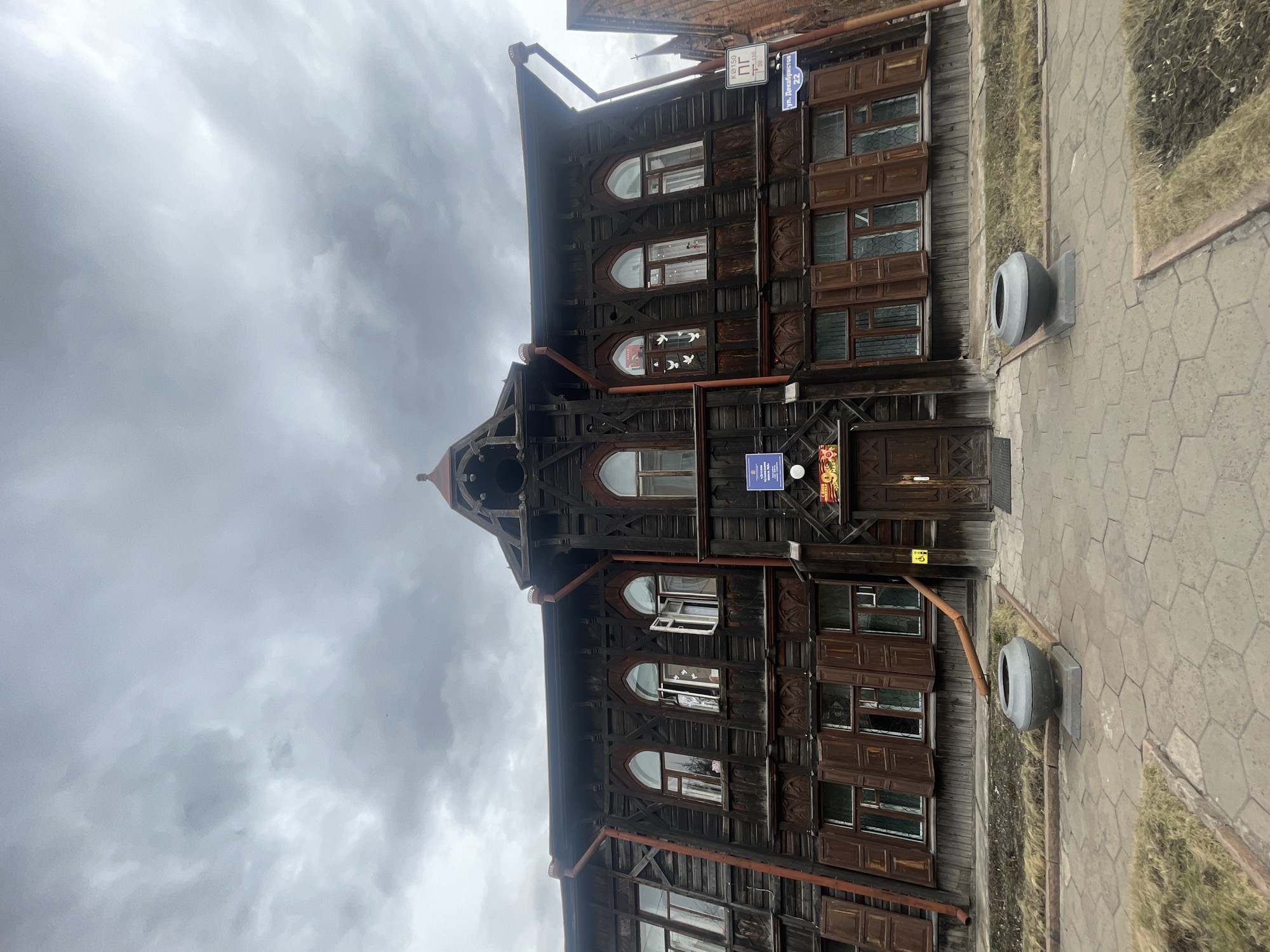 This media file captures a multi-story, dark brown wooden building with intricate carvings, located in Krasnoyarsk, Russia. The building's architecture features numerous windows, some with arched tops, and decorative wooden elements on its facade. A dark brown pipe runs along the exterior.
The scene is set under a heavily overcast sky, suggesting cloudy weather during the daytime. In the foreground, a paved sidewalk with hexagonal patterns is visible, alongside a narrow strip of dry grass. Two dark grey, urn-shaped trash cans or planters are situated on the sidewalk.
Several signs provide specific location details. A blue street sign clearly reads "ул. Декабристов 22" (Decembrists Street 22). Above it, a white sign shows "К.Э.150" and "21/10". On the building itself, a prominent blue sign identifies the establishment as "МБУК 'ЦБС' г. Красноярска Отдел 'Центр досуга и творчества' Библиотека № 15" (Municipal Budgetary Institution of Culture 'Centralized Library System' of Krasnoyarsk, Department 'Leisure and Creativity Center', Library No. 15), indicating it is a library. Another yellow sign displays "КРАСНОЯРСК" at the top with a red "50" on a round background.
There are no people or specific activities taking place, presenting a static, architectural street scene. The overall impression is calm and somewhat somber due to the subdued lighting from the cloudy sky.