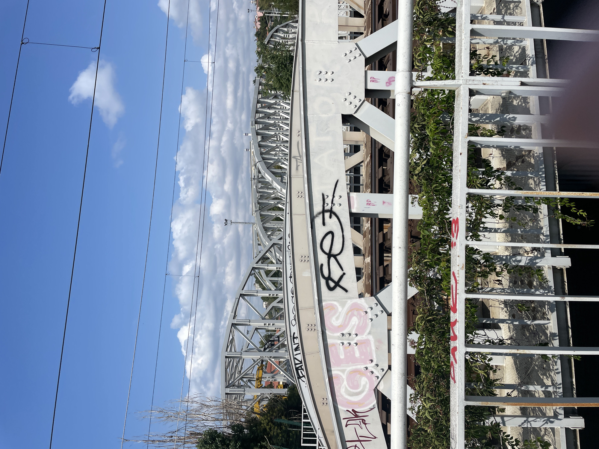 The image captures a view of a railway bridge in Kadıköy, Turkey. The bridge is constructed with light-colored metal beams and is adorned with graffiti. One section of the bridge features pink graffiti spelling out "GES" and a black stylized tag. Another part of the bridge has a black graffiti tag that appears to read "FAKIN." In the foreground, a white metal fence runs horizontally across the frame, partially obscuring the view of the bridge and the greenery growing behind it. The sky above is blue with scattered white clouds, and power lines are visible crisscrossing the upper portion of the image. The overall scene suggests an urban environment with infrastructure and elements of street art.