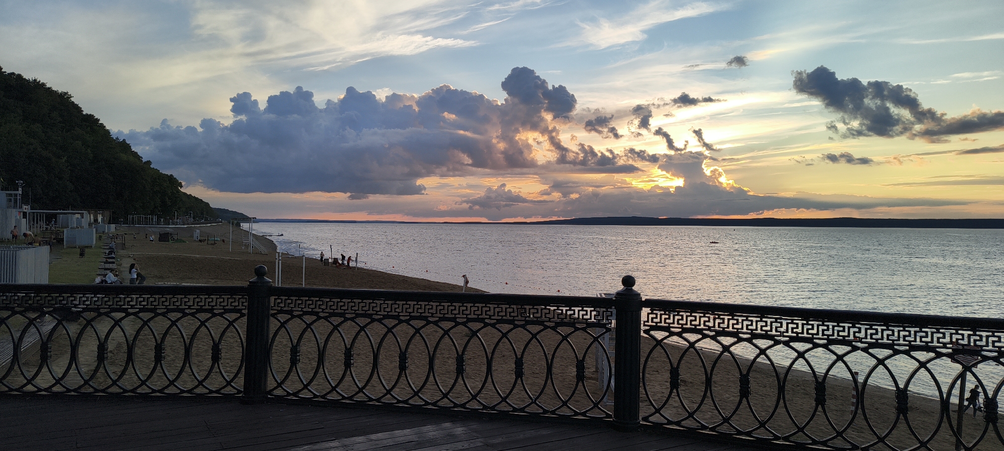 The image shows a wide, calm body of water at sunset. The sky is filled with dramatic clouds, illuminated by the setting sun which is partially obscured. A dark, tree-covered hillside dominates the left side of the scene. In the foreground, a dark, ornate metal railing with a geometric pattern separates the viewer from a sandy beach area. The beach has several small structures and a few people scattered about. The overall atmosphere is peaceful and serene, with the vibrant colors of the sunset reflecting on the water.