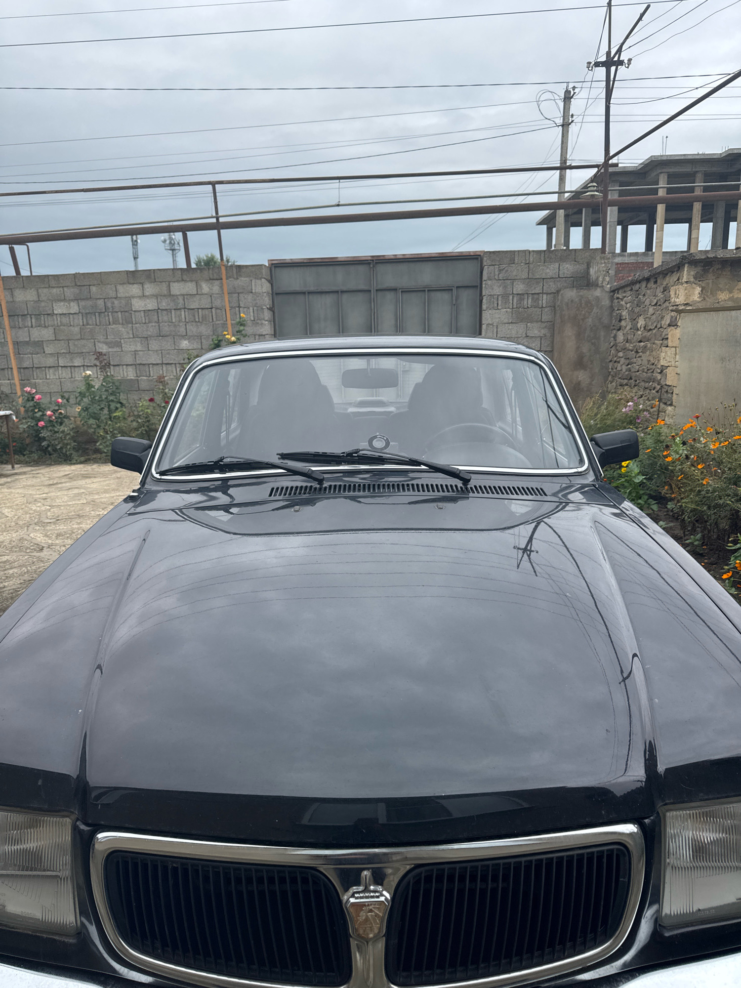 A dark-colored sedan is parked in an outdoor area, possibly a yard, with a cloudy sky overhead. The front of the car, including the hood, grille, and headlights, is clearly visible. Reflections of power lines and overhead structures are visible on the car's glossy surface. In the background, a grey brick wall and a stone wall are present, with some greenery and flowers to the left and right of the vehicle. A partially constructed building with concrete pillars can be seen in the distance. The image appears to have been taken from a low angle, looking slightly upwards at the car and its surroundings.