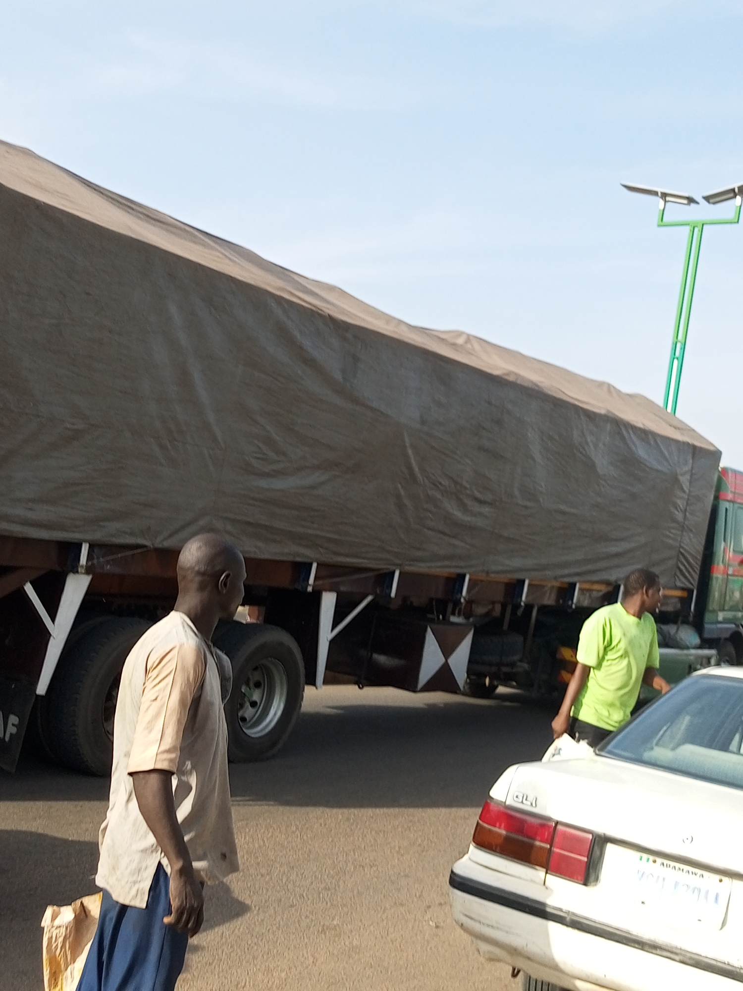 This media file depicts an outdoor scene, likely a road or street, during the daytime. The sun is shining, casting shadows and indicating clear weather. The primary focus is on a large, tarp-covered truck, suggesting it is loaded with goods for transport. Two men are visible, one standing near the front of the truck and another closer to the camera, walking towards a white car. The man near the truck is wearing a light-colored shirt and dark pants, while the man nearer the car is in a bright green t-shirt and dark pants. The presence of the truck and the car, along with the people, suggests a moment of transit or loading/unloading activity. A solar-powered street lamp is visible in the background, which could imply an urban or semi-urban setting.
Notable details include the fabric texture of the tarp covering the truck and the distinct pattern of the vehicle's tires. The white car has its taillights illuminated and a license plate visible, though the text is not entirely legible. The license plate has text that appears to read "ADAMAWA" and a series of numbers and letters below it, possibly "YGU 12011". The scene conveys a sense of everyday activity, possibly related to commerce or transportation. The expressions on the faces of the individuals are not clearly discernible, but their posture suggests they are engaged in their respective actions. The overall atmosphere is one of a functional, bustling environment.