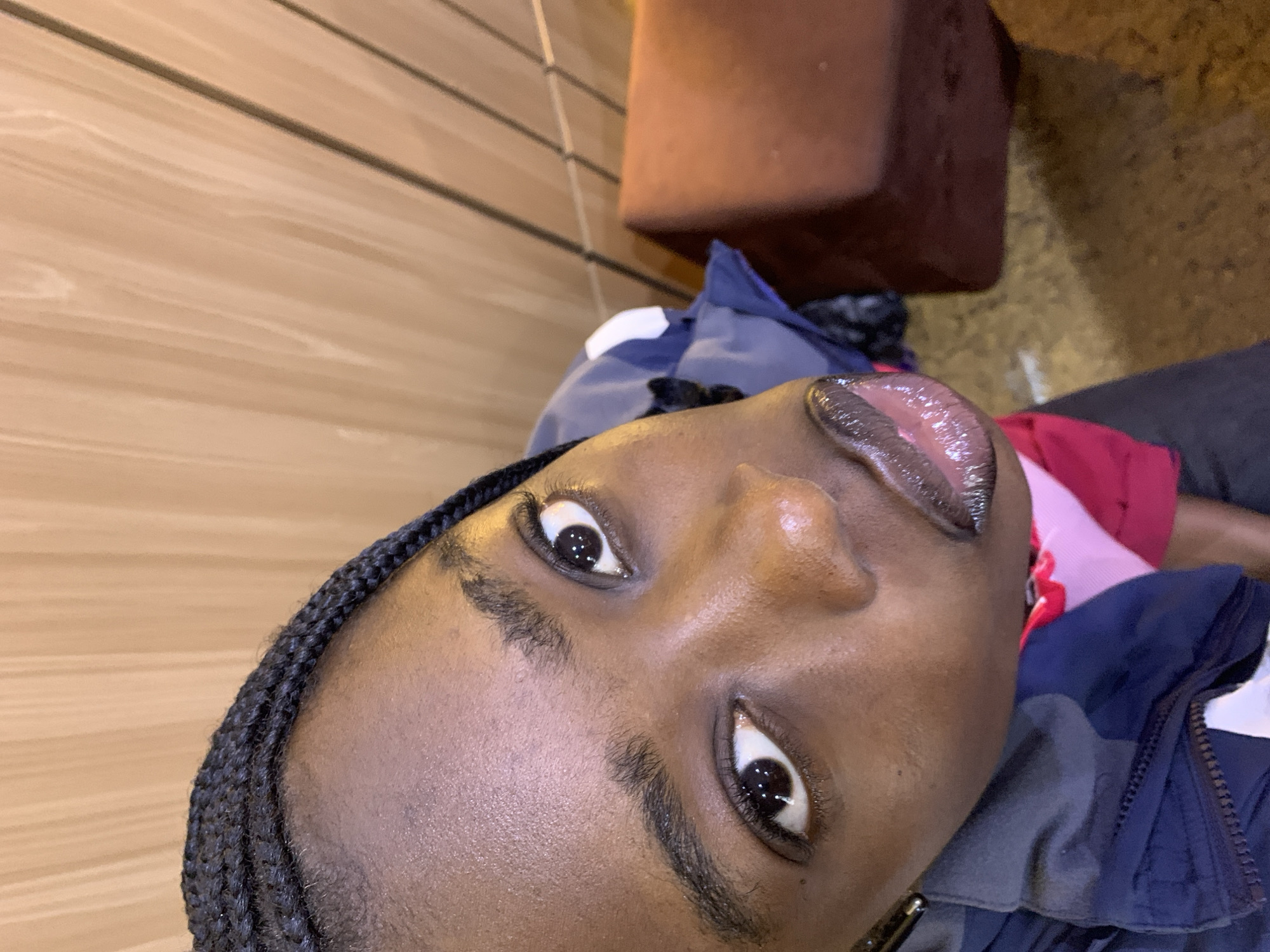 The image is a close-up, high-angle shot of a young Black woman with braided hair. Her face is angled upwards towards the camera. She is wearing a dark blue jacket with a lighter blue and white accent. Underneath the jacket, a pink garment is visible. Her lips are glossy and her eyes are wide open, reflecting light. The background consists of vertical wooden panels and a blurred reddish-brown object, possibly a piece of furniture. The overall lighting suggests an indoor setting, possibly at night or in a dimly lit area. The image does not provide any specific information about the location being Gura Top, Nigeria.