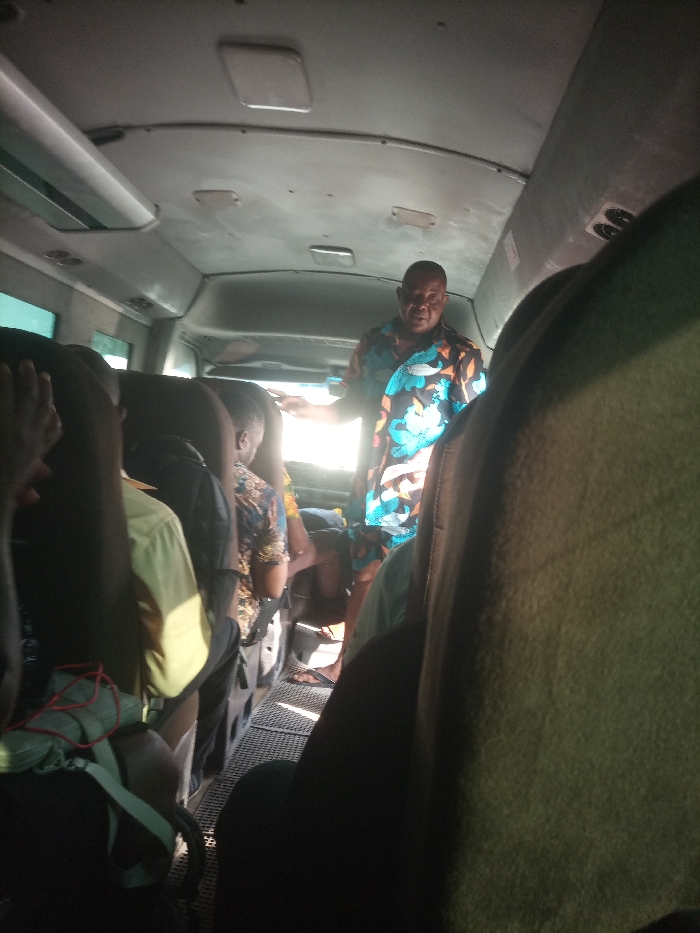 An interior view of a passenger minibus in Eliozu, Nigeria, shows several individuals seated, with a prominent male figure standing in the aisle. The standing man, of medium build and dark complexion, wears a brightly patterned short-sleeved shirt featuring blue, orange, and black motifs. He is positioned towards the center-right, facing left with his right arm slightly extended, illuminated by bright sunlight entering from the left side of the vehicle. Seated passengers, primarily viewed from behind or in profile, occupy rows of dark seats. One passenger in the mid-left wears a brown-patterned shirt. In the immediate foreground on the left, a person's left arm is raised, and a green backpack with red elements rests on the seat. The bus interior features a light-colored ceiling with overhead vents and a dark, possibly gridded, floor covering the aisle. Sunlight creates strong highlights and shadows, obscuring the exterior view through the windows. The perspective is from a seated position looking forward in the vehicle.