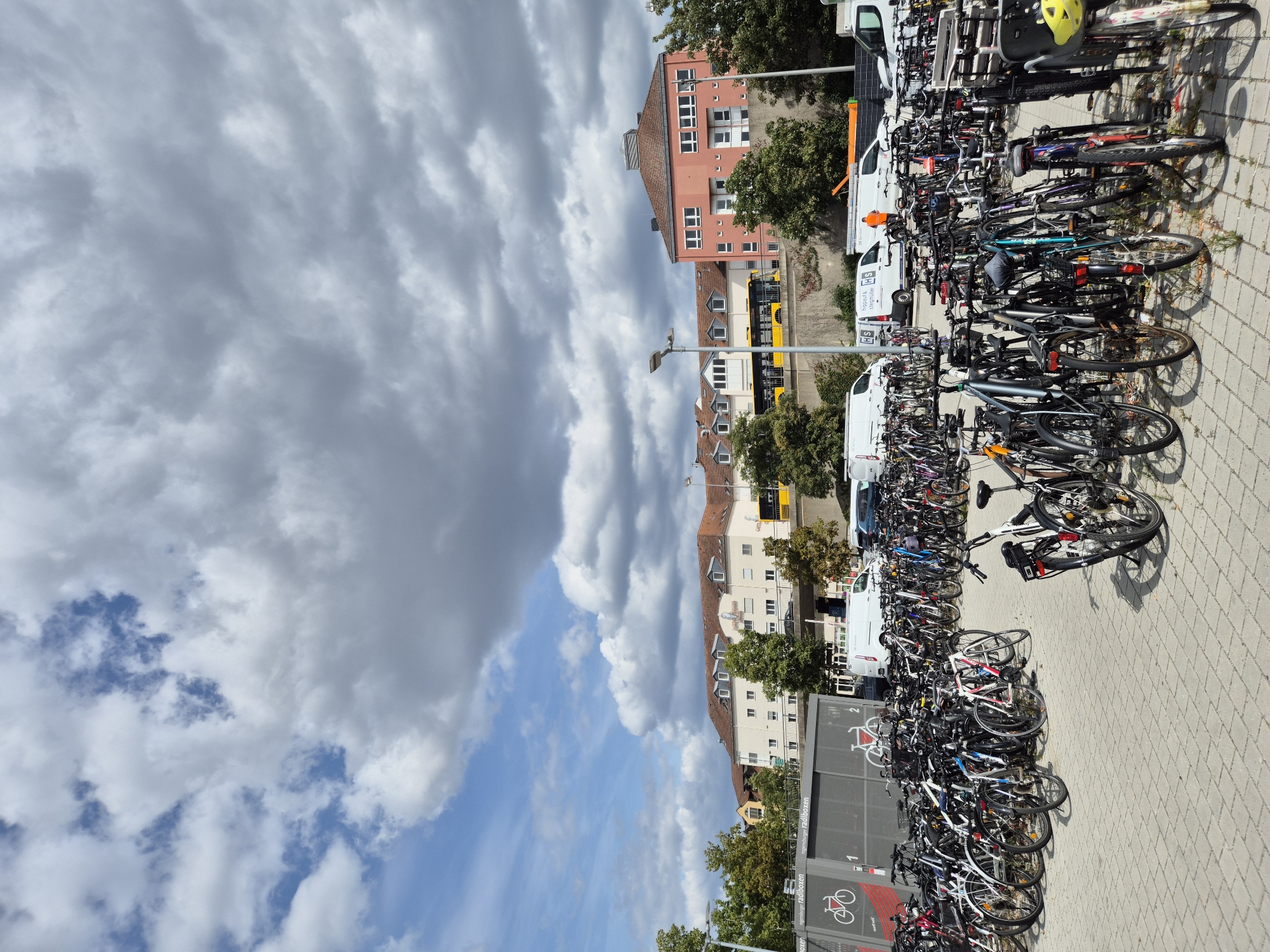 The image depicts a large outdoor bicycle parking area located in Regensburg, Germany. The scene is dominated by a vast number of parked bicycles of various types, neatly arranged in racks. The ground is paved with light-colored interlocking bricks. In the background, there are several multi-story buildings with tiled roofs, a yellow bus on an elevated road, and trees. The sky is filled with a mix of white and gray clouds. Several white vans are parked in the middle ground. There are no visible people actively engaged in actions within the frame, though the presence of bicycles suggests daily use by commuters and residents. The overall impression is that of a functional urban infrastructure designed to accommodate a significant number of bicycles.