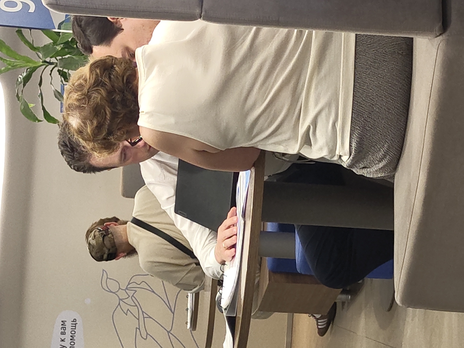 The image depicts an indoor scene, likely a cafe or co-working space, with several individuals engaged in work or discussion around a table. A person in the foreground, from the back, wears a cream-colored sleeveless top and grey textured pants. To their left, a man in a white shirt is looking down at a laptop and papers on the table. Another person, also from the back, wearing a tan sweater and a camouflage cap, is seated at the table. The environment includes a table with a laptop, papers, and notebooks. In the background, a plant and a blue sign with a white number "9" are visible. The presence of people working around a table suggests a collaborative or business-oriented setting. Without further context or distinctive landmarks, it is not possible to definitively determine if this location is in Ramenskoye, Russia.