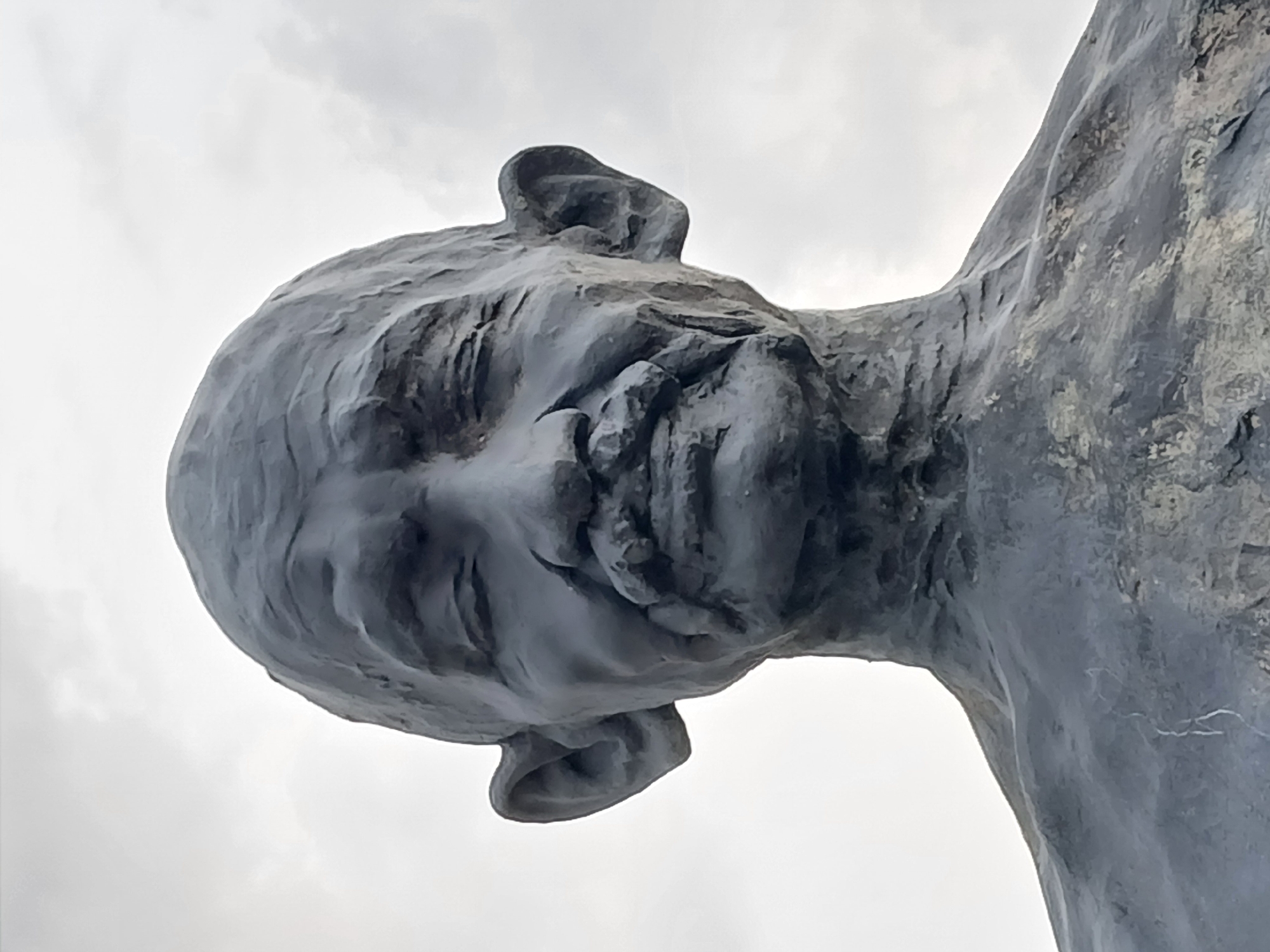 The image displays a large, dark-colored sculpture of a human head and the upper part of a torso, viewed from a dramatic low angle. The sculpture appears to be made of a textured material, possibly stone, concrete, or weathered metal, with a rugged, uneven surface. The human head is depicted with stylized features, including a visible ear, nose, and subtle contours suggesting closed or downcast eyes and a mouth. The head is tilted downwards or forward, creating a sense of contemplation or solemnity.
The sculpture dominates the foreground and midground, silhouetted against a bright, overcast sky. The sky is predominantly white and light grey, indicating a cloudy daytime setting. No other elements of the environment, such as ground, buildings, or landscape, are visible, making the sculpture appear monumental and isolated against the vastness of the sky. There is no discernible activity or event taking place, as it is a static art installation. No text is visible anywhere in the frame. This sculpture is located in Guayana City, Venezuela.
