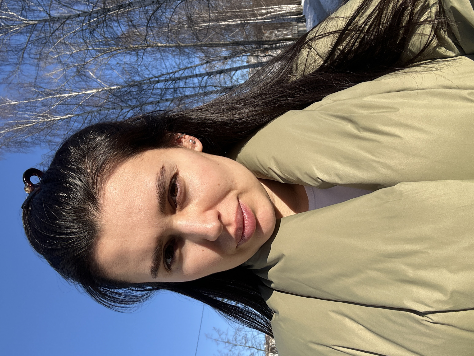 The image displays a close-up outdoor portrait of an adult female located in an area of Chaikovsky, Russia, characterized by bare trees and snow. The specific address within the city is not discernible from the visual information. The female subject has dark, straight, long hair, which is partially pulled back with a dark, possibly tortoiseshell-patterned, hair clip positioned near the top of her head. She has brown eyes and a neutral facial expression, with her lips slightly parted. Her right ear features multiple small, metallic stud and hoop piercings. She is wearing a prominent light olive green or sage green puffy jacket, with a white garment visible at the neckline beneath it.
The environment behind the subject consists of a clear, bright blue sky in the upper left, transitioning to an expanse of numerous bare deciduous trees, primarily birches, on the right side. The trees show light-colored bark. Patches of snow are visible on the ground beneath the trees, indicating a winter or early spring season. Faint outlines of multi-story buildings can be seen in the far background through the trees on the right. The scene is illuminated by direct sunlight, casting some shadows and highlighting the subject's face.