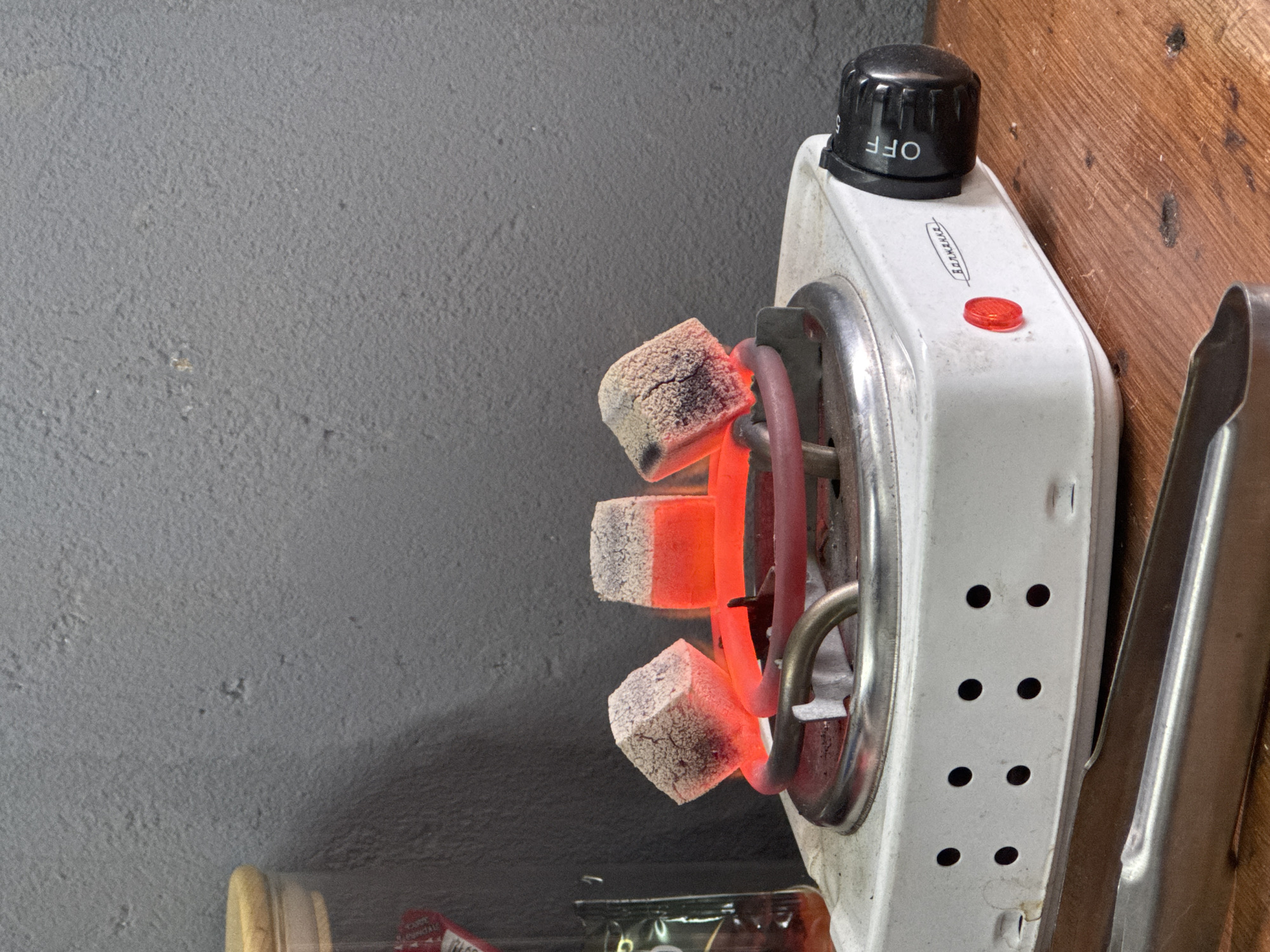 The image shows a white electric stove with a black knob labeled "OFF," with three charcoal briquettes glowing red on the heating element. The stove is next to a wooden wall. A pair of metal tongs is clipped to the side of the stove. The stove is set against a gray wall. A container is visible at the bottom left of the image. It's not possible to ascertain the location in Chaikovsky, Russia, from this image alone.