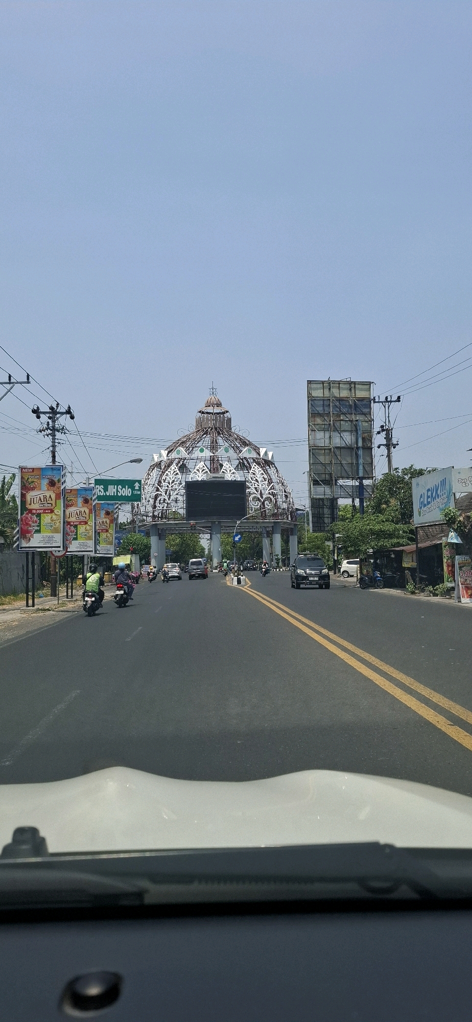 The image depicts a street scene in Surakarta, Indonesia, likely in the vicinity of the Gladak Plaza. In the foreground, the dashboard and windshield of a vehicle are visible. The road is asphalt with double yellow lines marking the lanes. Several vehicles, including cars and motorcycles, are on the road, with some motorcycles in the process of overtaking.
On the left side of the road, there are billboards advertising various products. One prominent billboard displays "JUARA" juice products, and another indicates "RS. JIH Solo 1.6 km". Further down the road, a large, ornate dome-like structure with intricate metalwork and a dark screen at its base dominates the view. This structure appears to be a landmark.
To the right of the dome, a tall, multi-story scaffolding structure is present, possibly for construction or advertising. Beyond that, a billboard with the text "GLEKK!!!" is visible, along with a small building that appears to be a shop or food stall. Trees and utility poles with wires are scattered throughout the scene. The sky is clear and blue, suggesting a sunny day. The overall impression is that of a busy urban street with significant traffic and commercial activity.