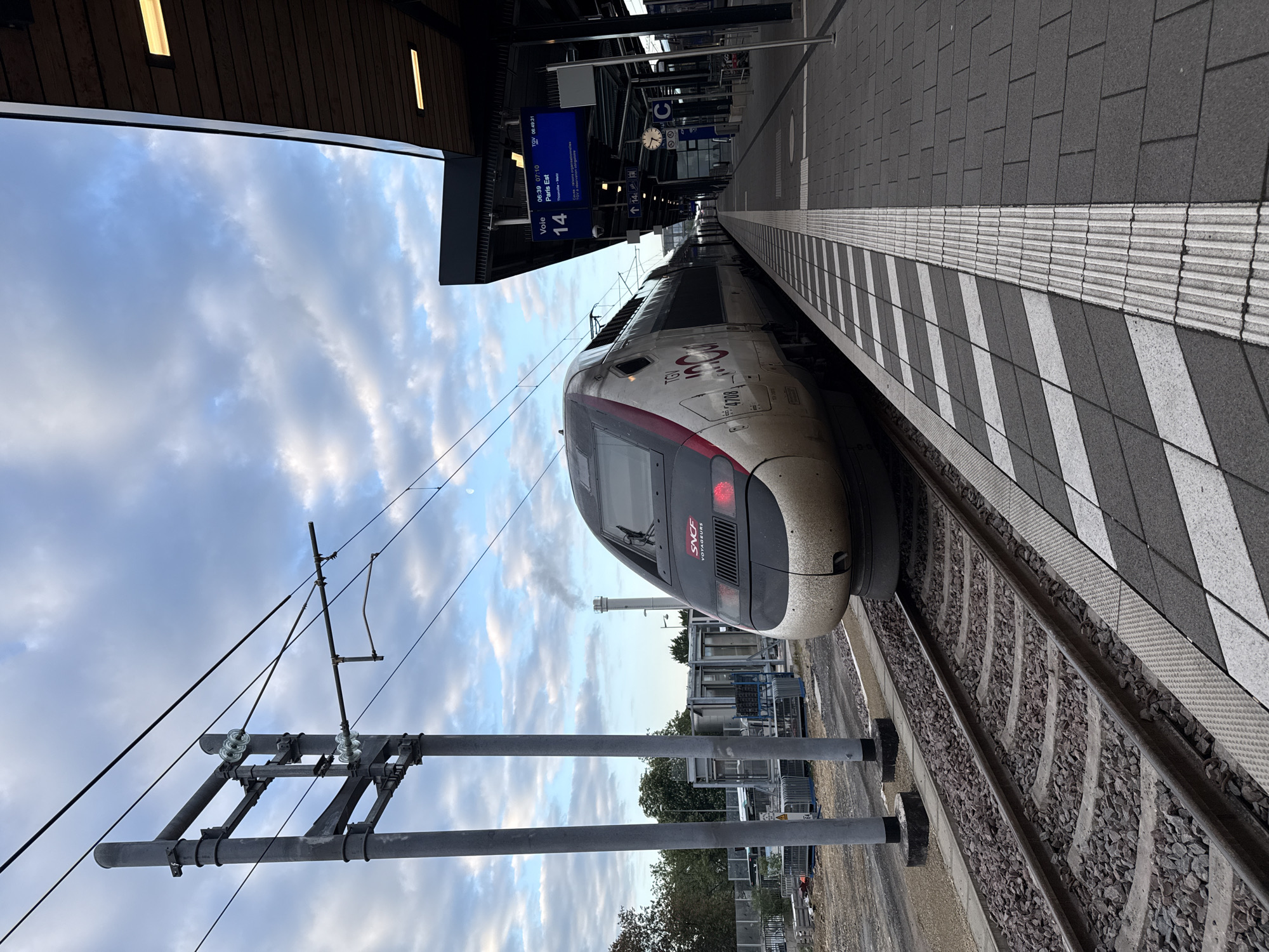 Here is a detailed description of the image:
**Location:** The image appears to be taken at a train station platform in the city of Luxembourg, Luxembourg.
**Subjects & Objects:**
* A high-speed TGV train is positioned at the platform. The train's front section features the SNCF logo.
* The platform has a distinct edge marked with a black and white striped safety zone.
* Overhead lines and supporting structures are visible.
* Signage indicates "Voie 14" (Track 14), along with departure information.
* Train station architecture is noticeable, including the platform canopy and the lighting fixtures.
**Environment:**
* The sky has a mix of blue and cloud cover.
* The platform surface has paving stones.
* The train tracks are filled with gravel.
* The weather appears to be daytime.