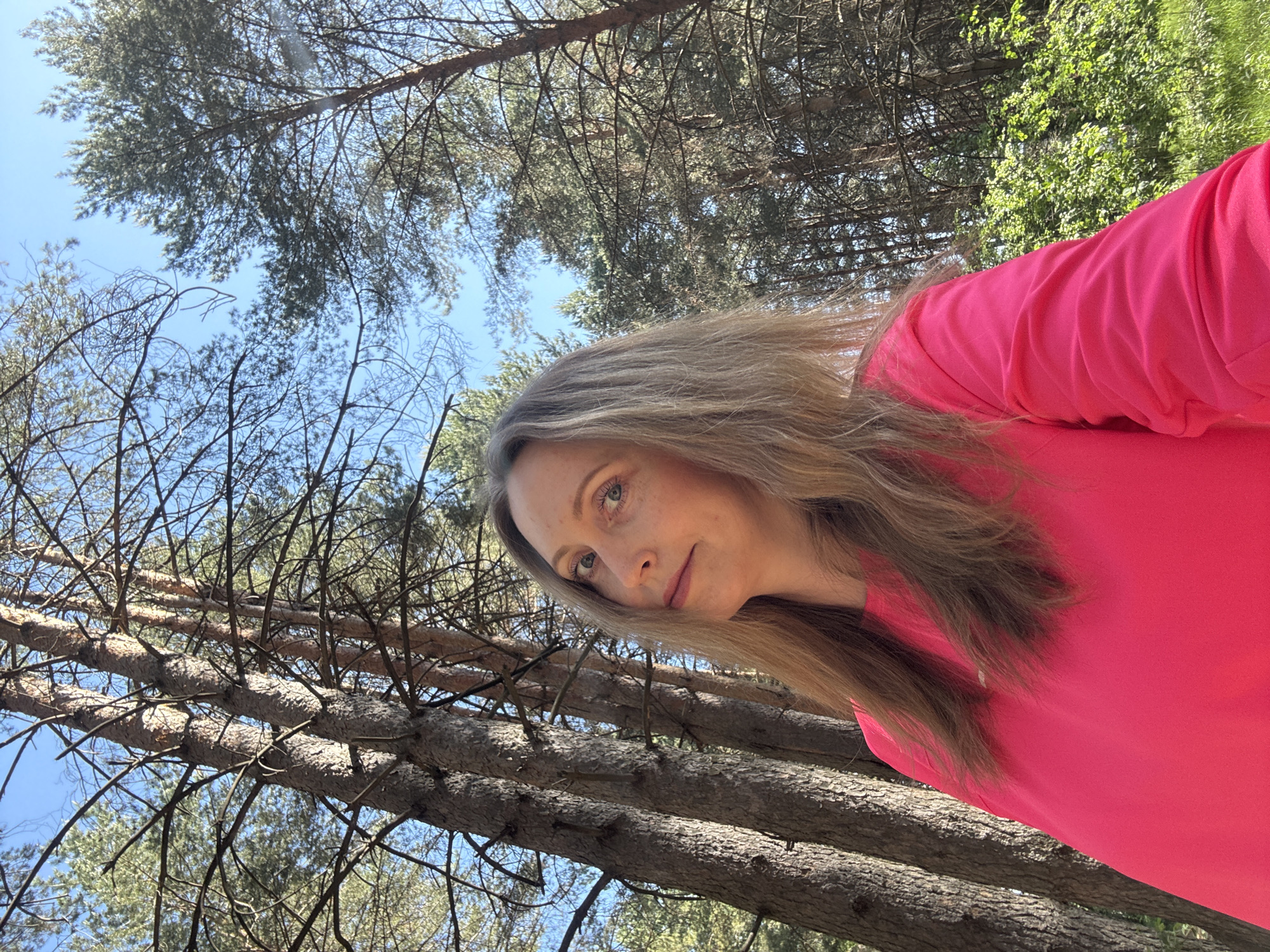 A woman is photographed outdoors in Chaikovsky, Russia. The woman has long, light brown hair, fair skin, and is wearing a bright pink long-sleeved shirt. The camera is positioned at an angle looking up at the woman, and the background is composed of trees and sky. The trees are mostly pine trees, with some deciduous trees mixed in. The sky is a light blue color. The photo appears to be taken on a sunny day in a wooded area, possibly a park.