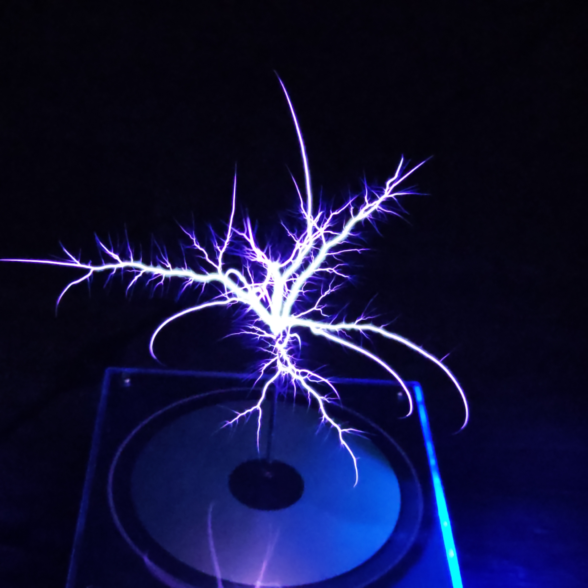 A close-up shot shows electrical discharges emanating from a device, possibly a Tesla coil. The discharges are a bright white-purple, branching out in a fractal pattern against a black background. The device sits on a dark, reflective surface. The image appears to have been taken indoors in low-light conditions. Location information is unavailable.