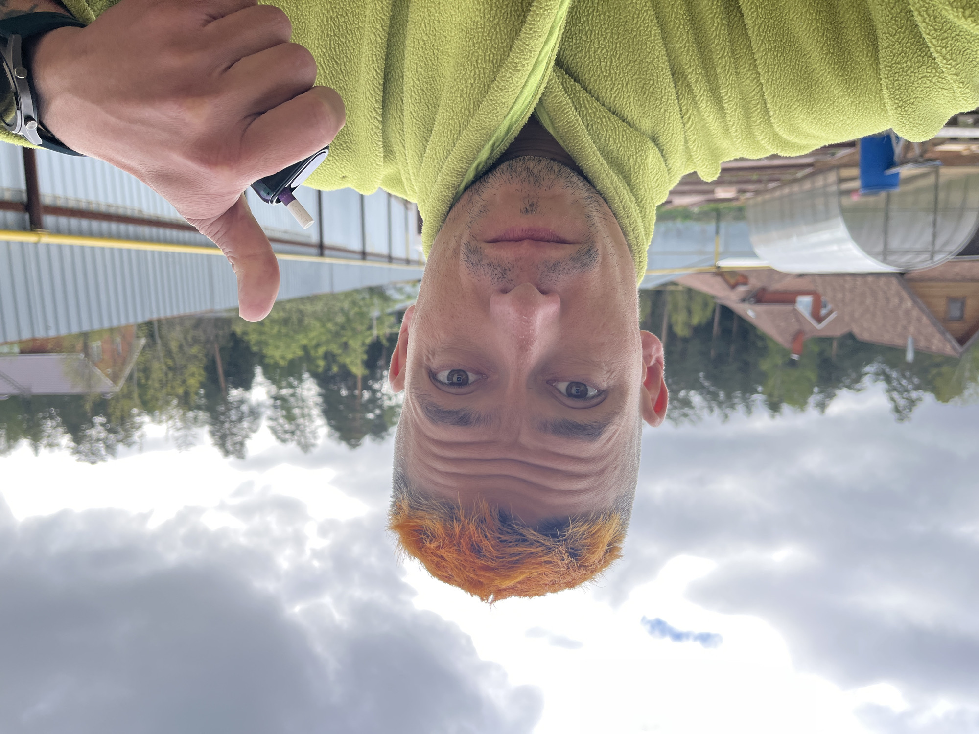 A man is holding a smoking device in his left hand, giving a thumbs down. He is wearing a lime green bathrobe. The man has brown eyes, a mustache, and a shaved head with orange dyed hair at the front. The sky is cloudy. In the background there is a house, a greenhouse, and trees. The location is Chaikovsky, Russia.