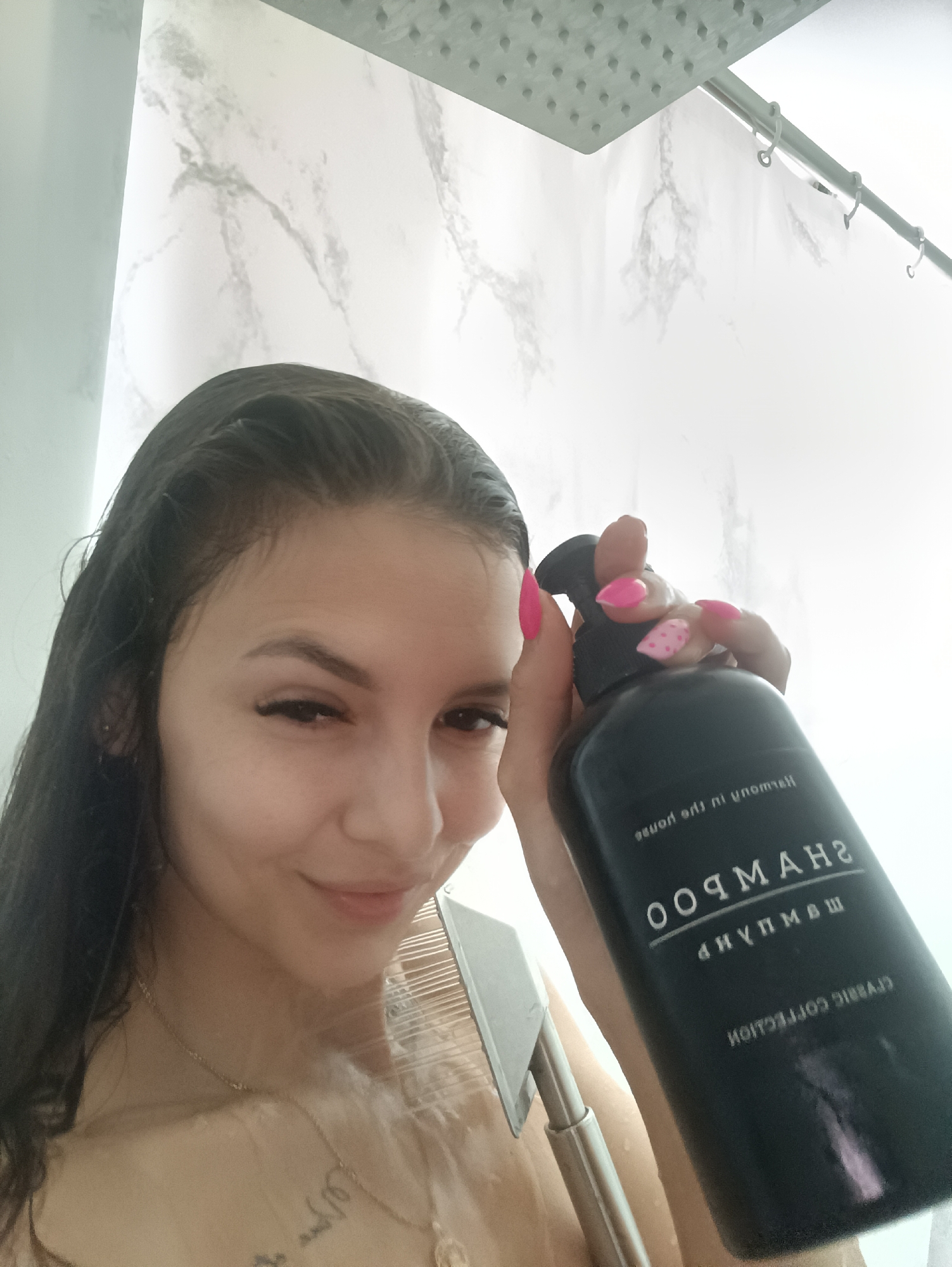 A young woman is in a shower, holding a black shampoo bottle with the word "SHAMPOO" written on it. Her wet hair is dark, and she has pink fingernails, one with polka dots. A gold necklace is visible around her neck. Water is running from a showerhead above, and a detachable shower head is also visible. The shower walls have a marble-like pattern. The location is unknown.