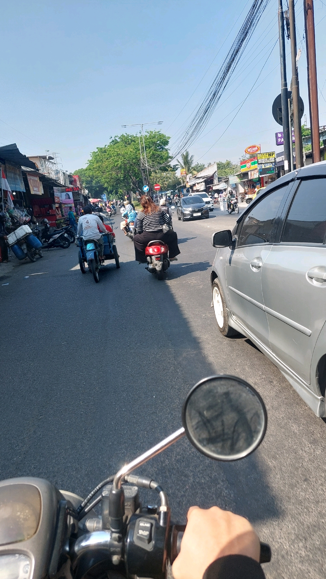 A first-person perspective shot from a motorcycle on a city street in Sidoarjo, Indonesia shows a busy road with traffic. A silver car is in the right lane, and a person on a scooter wearing a black and white striped shirt is in the left lane. Another person on a three-wheeled motorcycle is also in the left lane, carrying large blue containers. Shops and stalls line the left side of the road, with people walking and riding motorcycles. The sky is clear and blue, and trees are visible in the background. The motorcycle's handlebars and a hand gripping them are visible in the foreground.