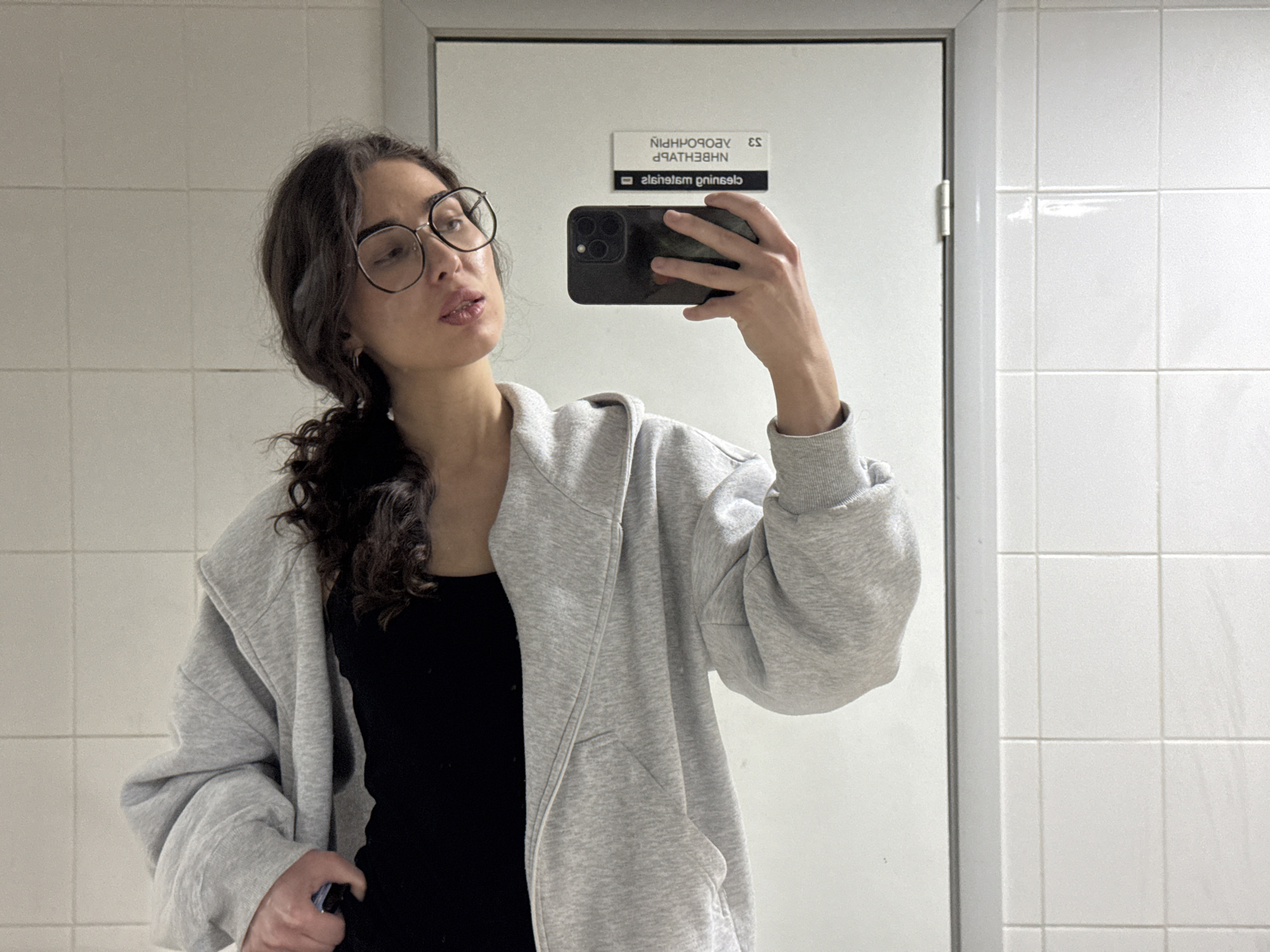 A young woman with dark, curly hair pulled back into a ponytail, wearing round glasses, a black tank top, and a grey zip-up hoodie, takes a mirror selfie in what appears to be a restroom. The environment is tiled in white on the right side of the frame. A sign is visible on the door behind her, with text that reads "CLEANING ESSENTIALS" and other text in Russian. The woman is holding a black smartphone to take the picture.