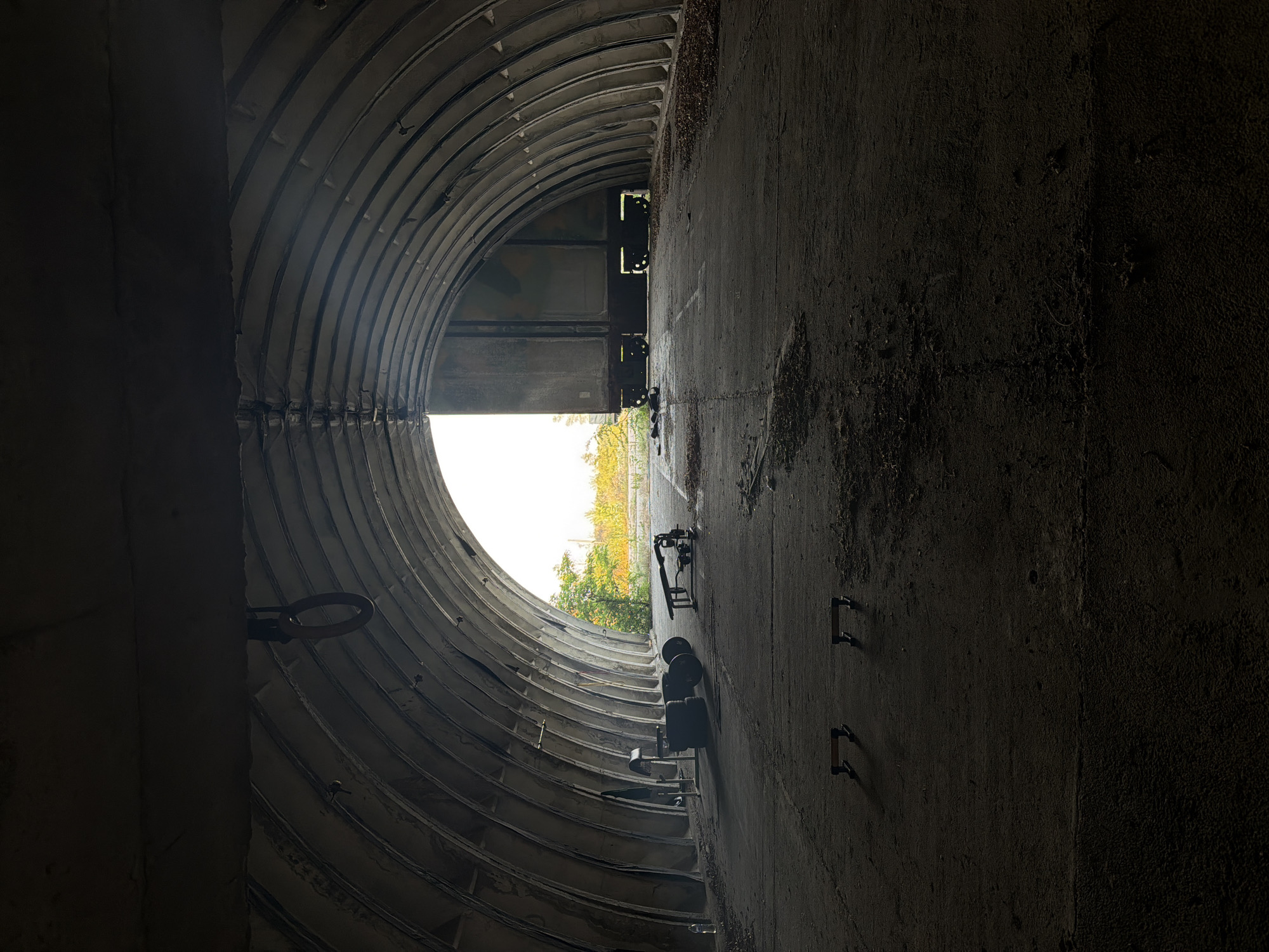 The image displays the interior of a large, arched structure, likely a bunker or hangar, constructed with visible corrugated metal or concrete ribs forming the ceiling. The perspective is from a dark interior towards a brightly illuminated exterior opening. The floor is a rough, unpaved concrete or dirt surface covered with scattered debris, including dried leaves and small particulates.
In the mid-ground on the left, several pieces of exercise equipment are arranged: a black upholstered weight bench, a stack of large, dark weight plates, and a dark towel or fabric draped over a metallic frame. A clear plastic water bottle is visible on the floor near the equipment. In the foreground, two dark push-up bars rest on the ground, casting distinct shadows to their left.
On the right side of the mid-ground, another weight bench is positioned, partially obscured, with additional exercise equipment nearby. A large, dark, rectangular structure, resembling a heavy door or a section of a railway car, is partially open at the main exit, revealing bright light from behind it and contributing to the stark contrast in illumination.
Beyond the open end of the structure, the exterior environment is brightly overexposed, showing dense green and yellow-orange foliage under what appears to be an overcast or bright sky. A single metal ring hangs from the ceiling support in the upper left portion of the interior. The scene is located in Bogdanitsa, Bulgaria.