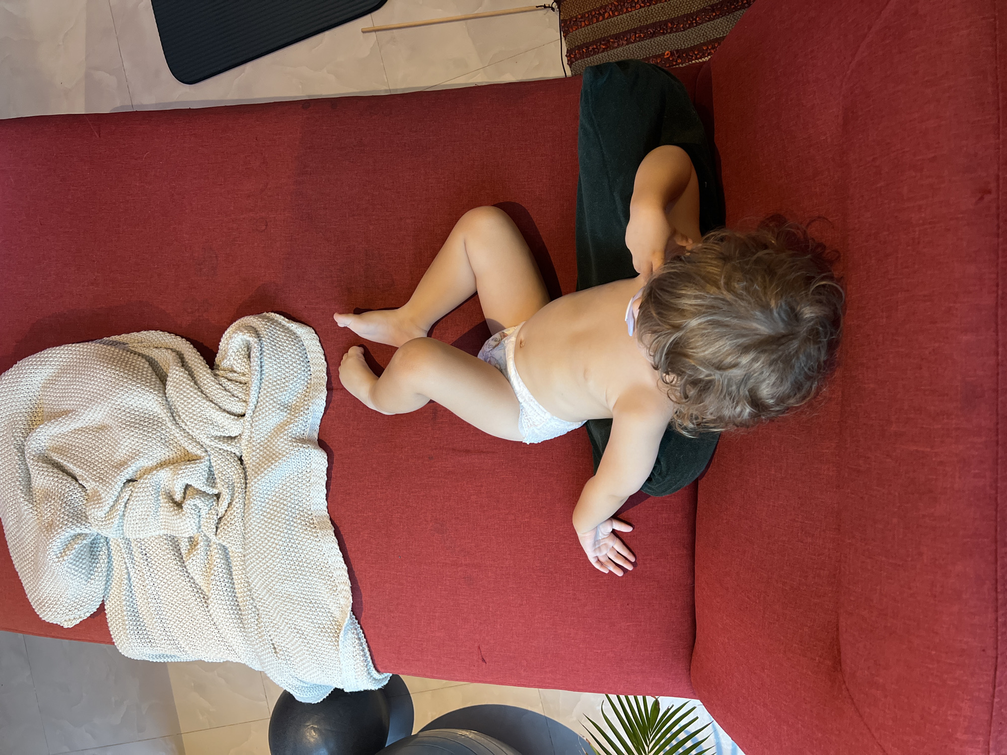 A high-angle shot captures a toddler lying on a red couch. The toddler, seen from the back and side, is wearing a diaper and has curly brown hair. Their body is mostly exposed, with one leg bent and the other extended. A white knitted blanket is draped over the left side of the couch, partially covering the toddler's legs. To the right of the toddler, a dark green cushion or pillow is visible. The scene appears to be indoors, with a tiled floor and a portion of a yoga mat visible in the upper left corner. Other objects on the floor, including exercise balls and a plant, are partially visible at the bottom. The lighting suggests it could be daytime.