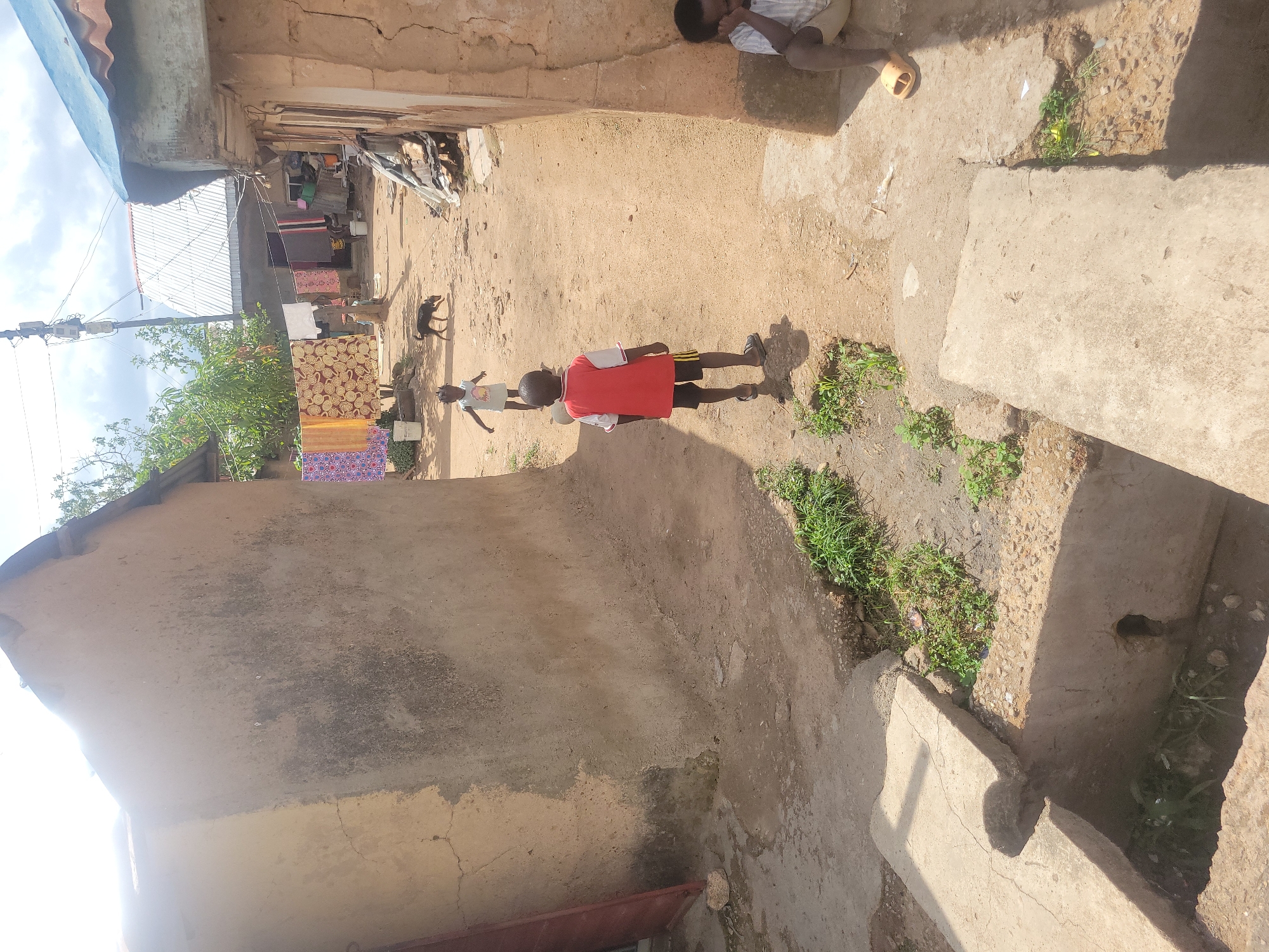 The image depicts an outdoor scene in what appears to be a residential area, possibly in Jos, Nigeria, though this location is not definitively confirmed by visual elements in the picture alone. The environment includes buildings with weathered walls and corrugated metal roofs.
Two children are visible in the center of the frame. One child is wearing a red shirt and dark shorts, while the other is wearing a lighter colored shirt. They appear to be playing or walking. A dog is also present, moving in the same direction as the children. Laundry is hanging on a clothesline in the background. Another individual is partially visible at the top-right of the frame.
The ground is primarily dirt, with patches of grass and weeds growing in certain areas. The presence of concrete structures and steps suggests some degree of development.