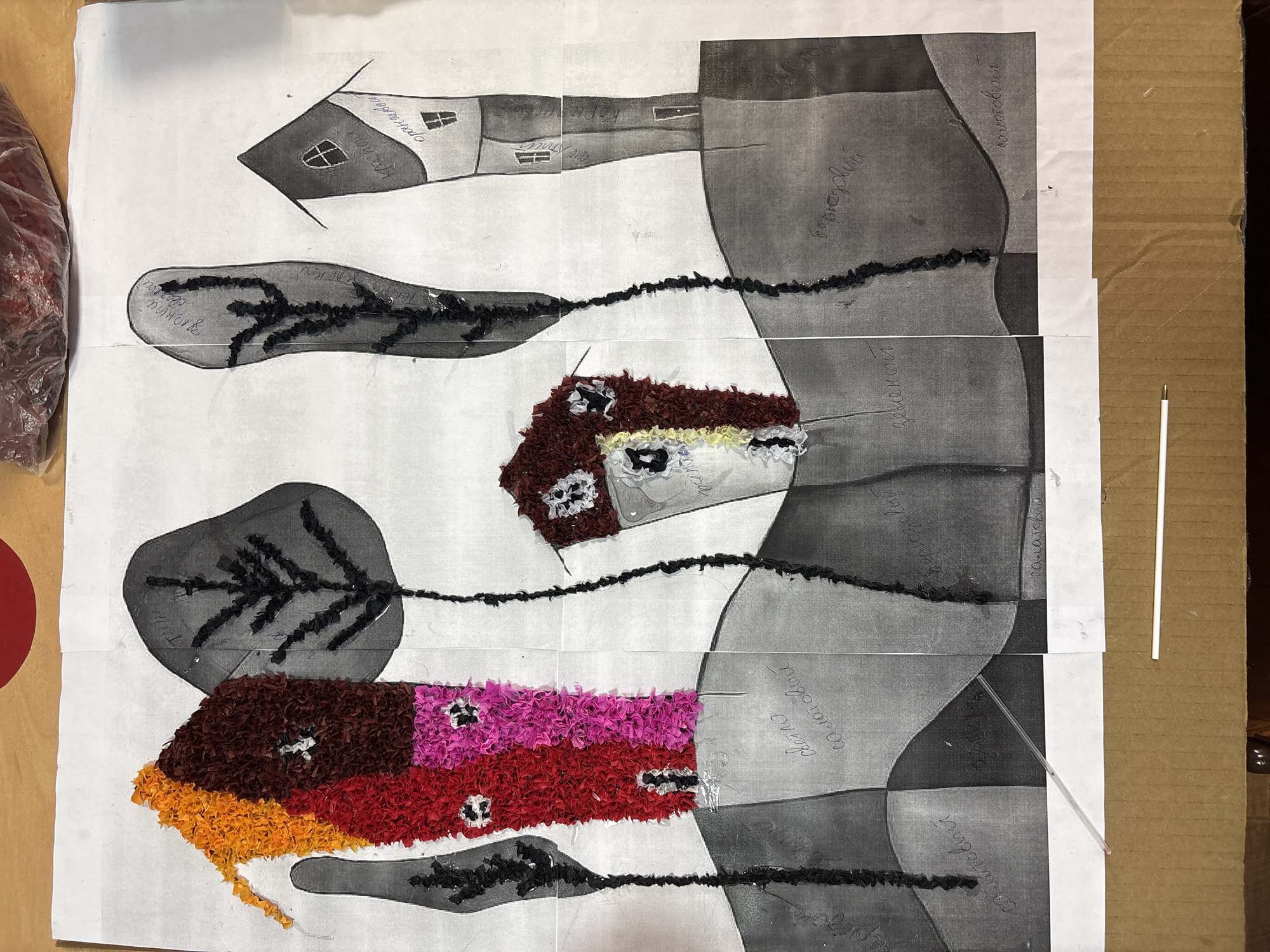 This is a collage artwork depicting a whimsical village scene, likely created by children. The artwork features several stylized houses made of colorful shredded paper, including houses in bright red, pink, and orange. These houses are interspersed with black, twig-like trees and set against a background of grayscale abstract shapes that suggest rolling hills or landforms. Some of the houses have small, dark windows. Inscriptions in what appears to be Cyrillic script are present on some of the grayscale elements, possibly labeling colors or elements of the scene. The artwork is mounted on paper and appears to be a composite of several sheets. In the foreground, a portion of a cardboard box is visible, and two white sticks, possibly straws or similar items, are laid across the bottom of the artwork. There are no people or animals visible in the image. The artwork is likely located in a home or classroom setting in Ветрынскі сельскі Савет, Belarus, given the presence of Cyrillic script and common artistic materials.