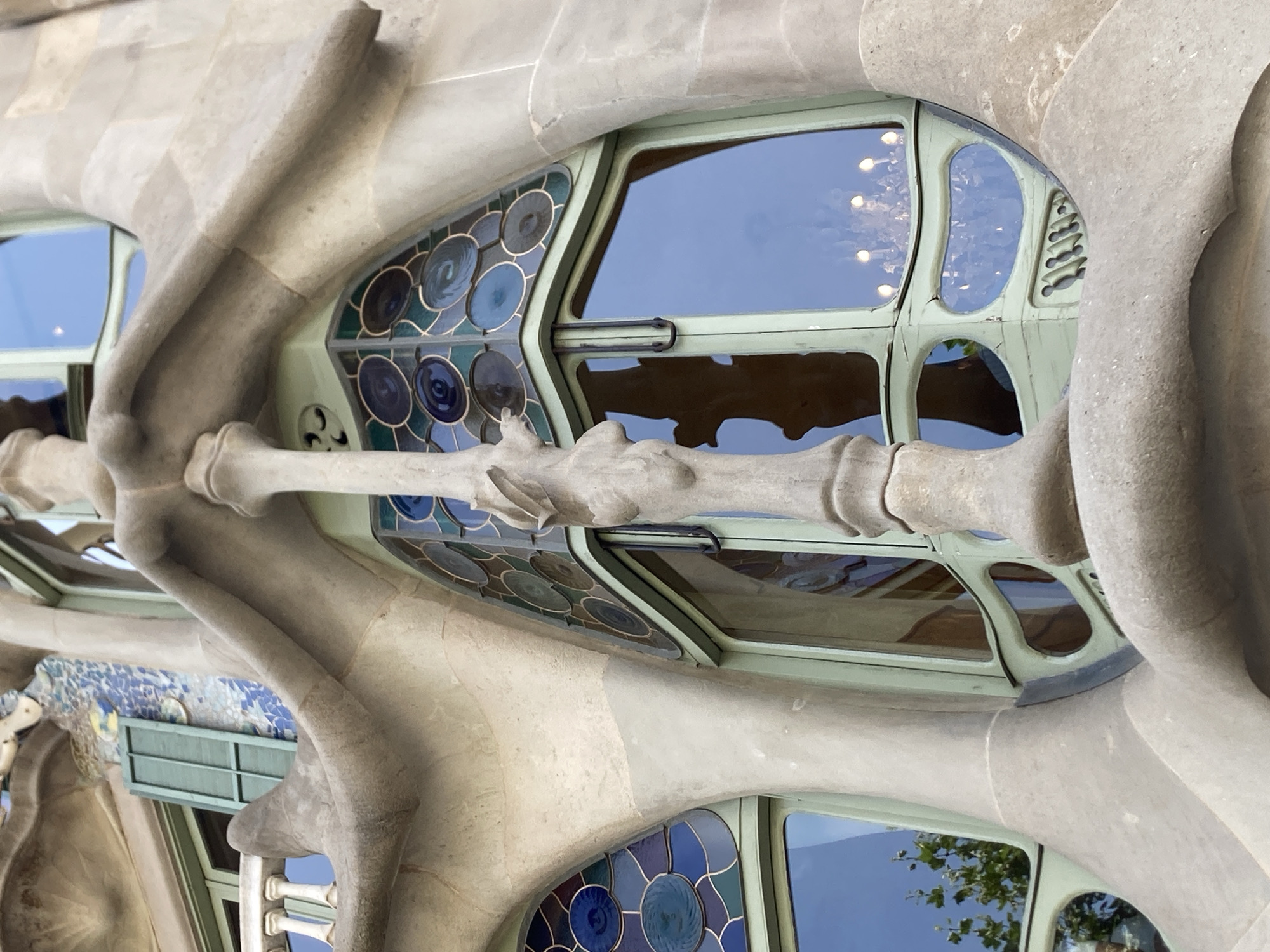 The image is a close-up, low-angle shot of the facade of Casa Batlló, a building located on Passeig de Gràcia in the Eixample district of Barcelona, Spain. The facade is characterized by its organic, flowing architectural style, which is typical of Antoni Gaudí's work. The photograph focuses on a section of the building featuring a large, irregular-shaped window with a distinctive pale green frame. The window is composed of clear glass panes that reflect the blue sky and surrounding architectural elements. Above the clear glass, there is a decorative section of stained glass with circular motifs in shades of blue and green, bordered by the same pale green frame.
A prominent, bone-like, light-colored stone column or structural element curves diagonally across the center of the frame, obscuring parts of the window and the underlying facade. This element appears to be a continuation of the building's fluid design, with a rough, textured surface. To the left of the main window, another similar window is visible, also framed in pale green and featuring stained glass with circular blue and darker circular patterns. Further to the left, a portion of a white balcony railing with vertical supports can be seen, as well as a green shutter and mosaic tiling on the upper part of the facade. The overall impression is one of a highly sculptural and unconventional architectural design.
