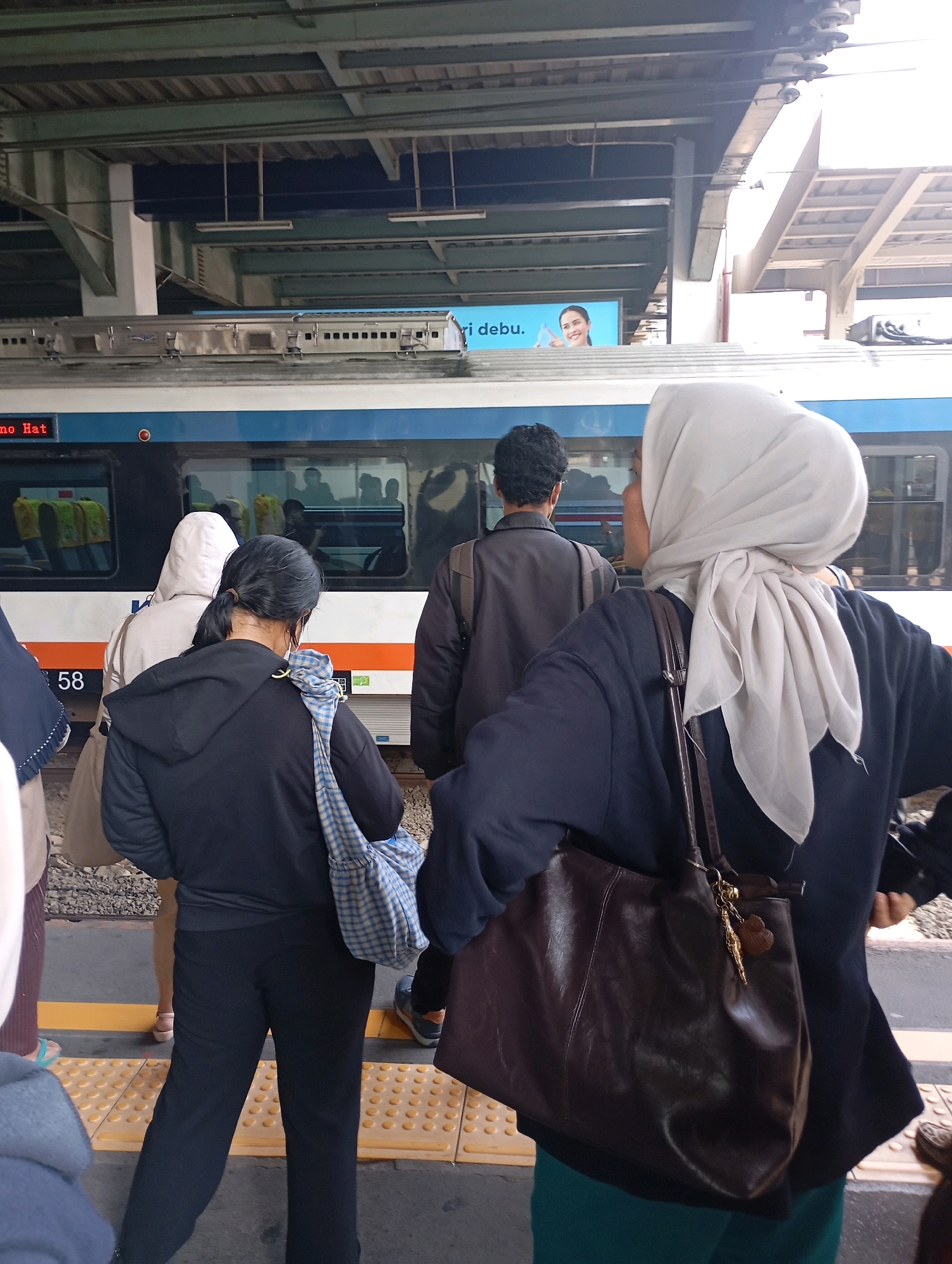 A low-angle, medium shot captures a group of people waiting at a train station platform in West Jakarta, Indonesia. In the foreground, a woman wearing a dark blue sweater and a light grey hijab is seen from behind, carrying a large brown leather bag. To her left, another person in a dark hoodie faces away from the camera. Further left, a person in a light-colored hoodie stands near a train with blue and white stripes. The train has windows through which several passengers are visible. The platform features yellow tactile paving and a metallic grey railing. In the background, the train station's elevated structure with metal beams and ceiling panels is visible, along with an advertisement featuring a woman. The lighting suggests it is daytime, possibly late morning or early afternoon, with sunlight casting some shadows.