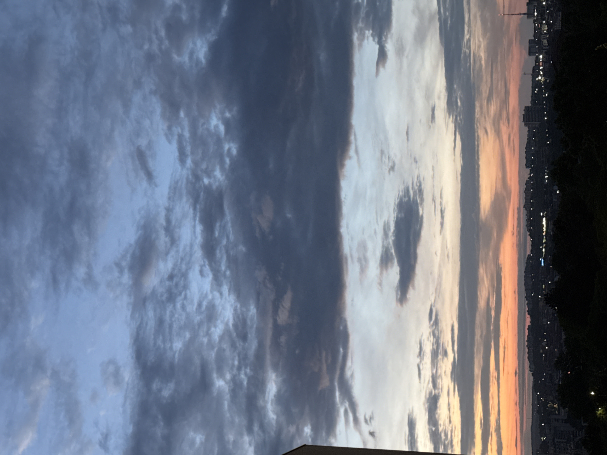 A cityscape is visible under a dramatic sky filled with dark, scattered clouds. The horizon is marked by a silhouetted line of buildings and trees, with scattered lights indicating an urban area. The sky transitions from a pale blue at the top to warmer hues of orange and pink near the horizon, suggesting either sunrise or sunset. The image appears to be taken from an elevated viewpoint, possibly a building or hill overlooking the city. There are no people or active interactions depicted in the scene. The overall atmosphere is serene and visually striking due to the contrast between the dark clouds and the soft colors of the sky. This scene could be interpreted as a view over Pleven, Bulgaria, given the context provided.