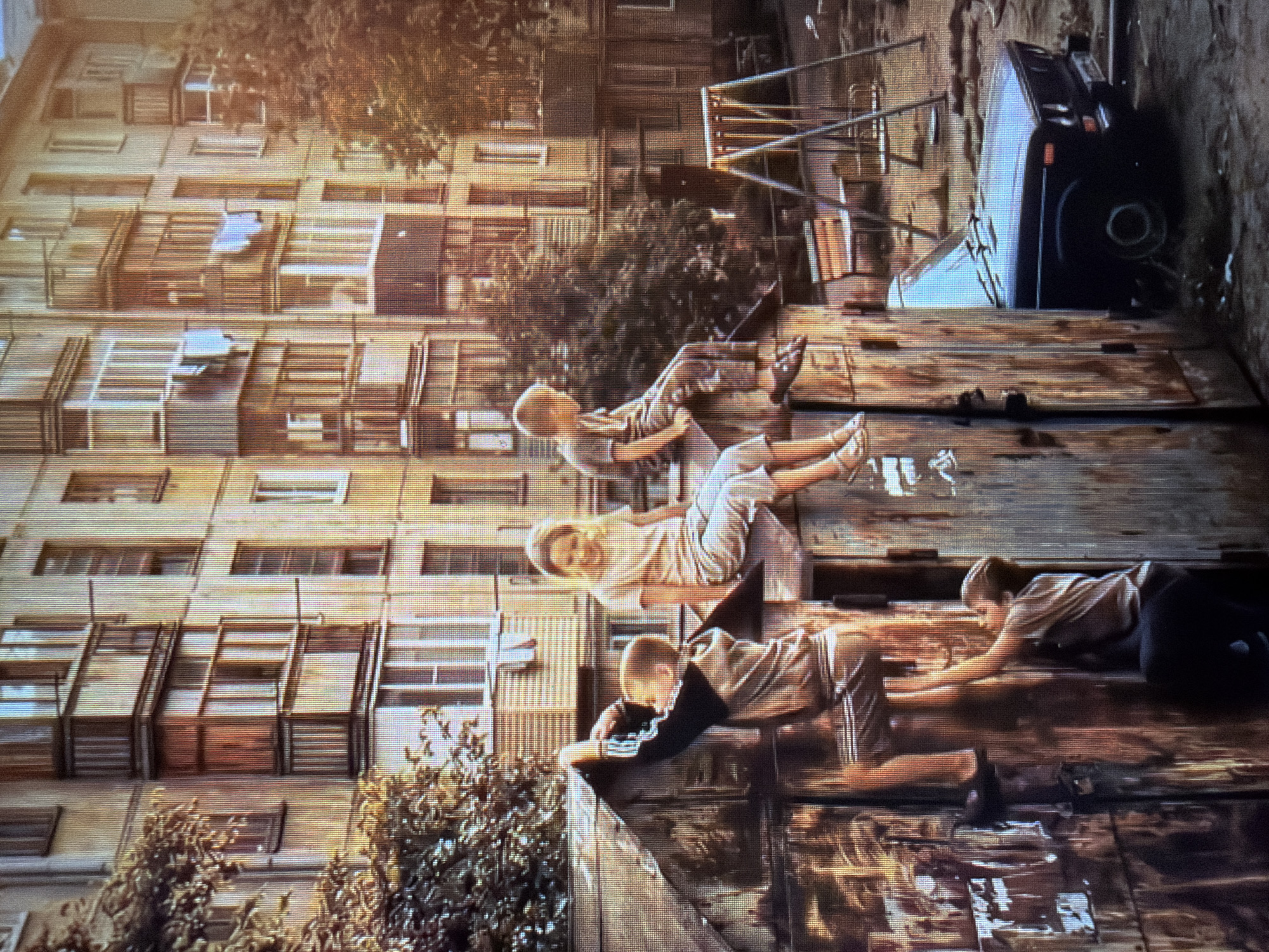 The image depicts a surreal scene with four young people in mid-air, appearing to be jumping or falling against the backdrop of a Brutalist apartment building. The building, with its repetitive windows and balconies, suggests an urban environment, possibly Eastern Europe. The people are positioned at different heights, their bodies contorted as if in freefall.
The first person from the top, a young girl with blonde hair, is seen floating horizontally with her legs extended. Below her, a boy with blonde hair is in a similar pose, his arms outstretched. Further down, another young girl with blonde hair is captured in a dynamic pose, her body twisted. The lowest person, a young boy, is shown reaching out, his body angled as if trying to grab onto something. Their clothing is casual and appears to be from the late 20th or early 21st century.
The ground beneath them is a weathered wooden surface, made of planks, which adds to the unusual and staged nature of the scene. In the background, there's a glimpse of a playground structure and a dark, angular object that might be a vehicle. The lighting suggests it is daytime, with warm tones hinting at either sunrise or sunset, casting a soft glow on the scene. The overall atmosphere is one of playful defiance of gravity, with a touch of mystery due to the unexplainable nature of their levitation. No text is visible in the image.