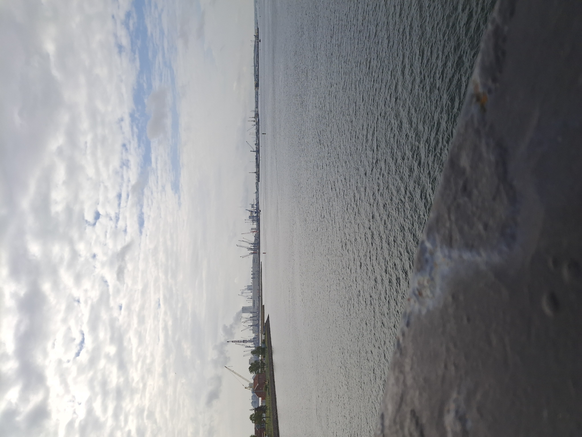 The image shows a view of the Rostock harbor in Germany. The photo appears to have been taken from an elevated position, potentially from a bridge or high structure. In the background is a cloudy sky. The water is visible in the foreground, with slight ripples. On the horizon line, there are cranes and industrial structures, typical of a busy port area. Some buildings are also visible near the shoreline. The perspective is slightly tilted. In the bottom right corner is a portion of a structure that seems to be made of gray concrete.