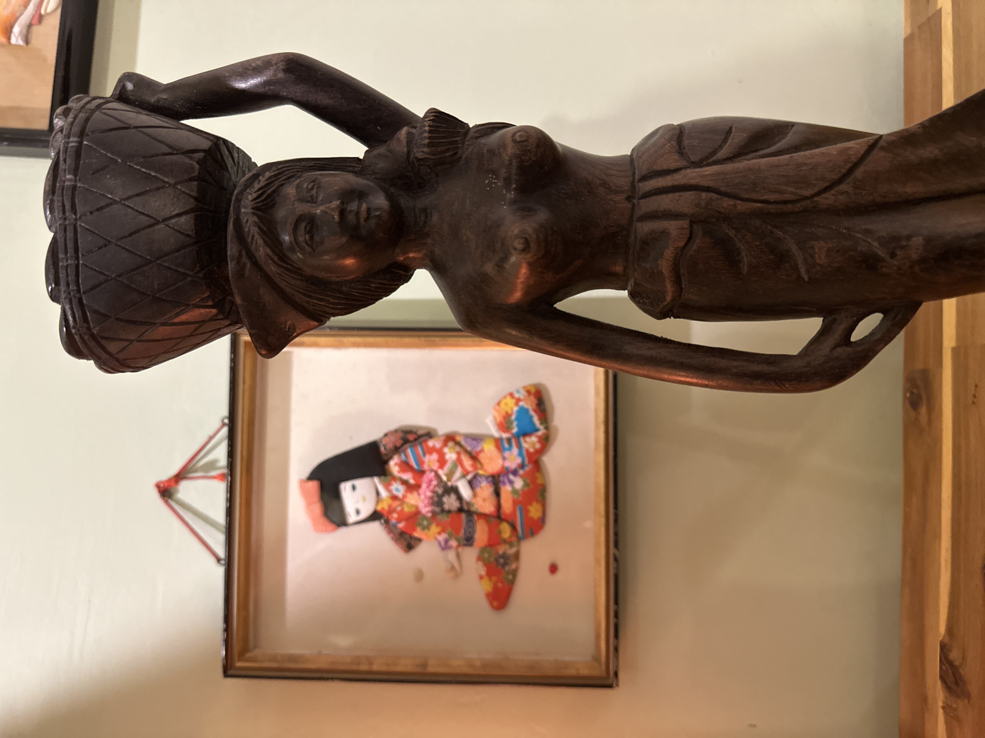 A dark brown wooden figurine of a woman carrying a basket on her head stands on a wooden surface in the foreground, slightly to the right. The figurine is carved in a somewhat stylized manner, with details suggesting a sarong around her waist and breasts. Behind the figurine and to the left, a framed artwork depicts a doll dressed in a colorful kimono, kneeling. The artwork is hung by a red string attached to a triangular metal bracket on the wall. The wall itself is a pale, muted green. The overall setting appears to be indoors, possibly a home or shop, given the decorative nature of the objects. No specific location in ตำบลเกาะพะงัน, Thailand can be determined from the image alone.