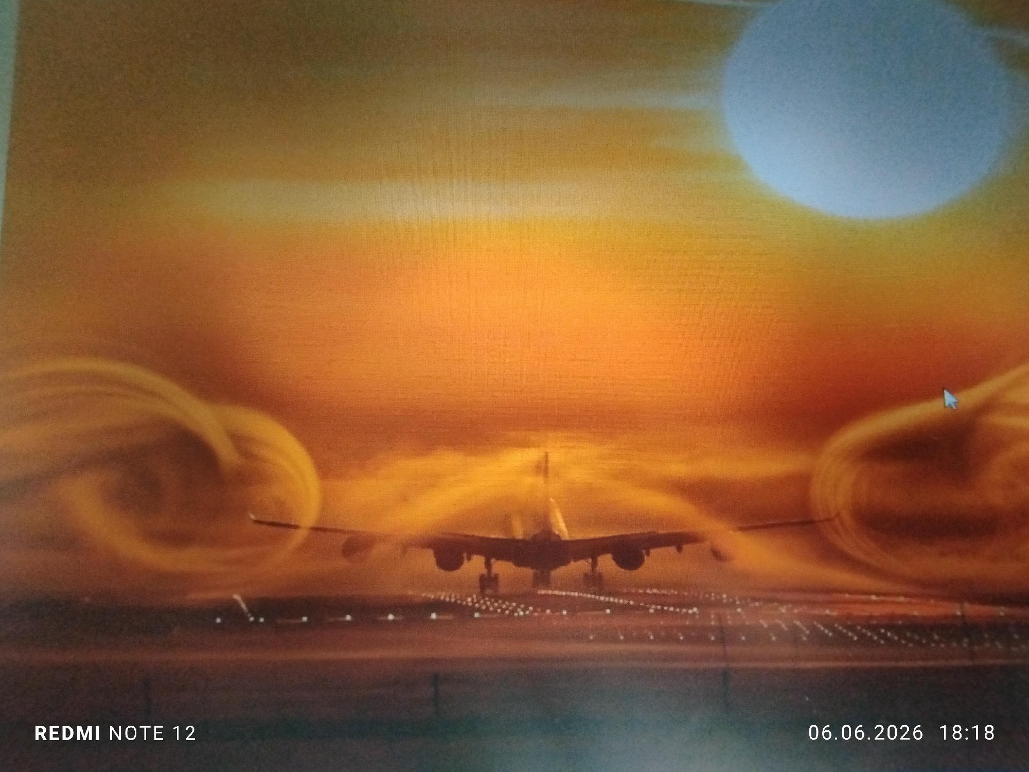 The image displays a large passenger aircraft positioned centrally on an illuminated airport runway, viewed from a rear perspective. The aircraft's landing gear is extended, indicating either a landing or takeoff maneuver. The environment is dominated by an intensely hued sky featuring a gradient of orange and yellow, characteristic of sunrise or sunset. A large, diffuse, pale blue-white circular object, resembling the sun, is visible in the upper right quadrant of the sky. Swirling orange and yellow atmospheric effects, appearing as vortices or jet wash, are prominent on both sides of the aircraft, emanating from the wingtip areas. The runway is delineated by multiple parallel lines of bright white lights extending into the distance. No individuals are present in the scene. A computer mouse cursor is visible in the upper right portion of the sky. Text overlays include "REDMI NOTE 12" in the bottom left corner and "06.06.2026 18:18" in the bottom right corner. The image contains no specific visual markers or identifiers to confirm its location as Burgas, Bulgaria.