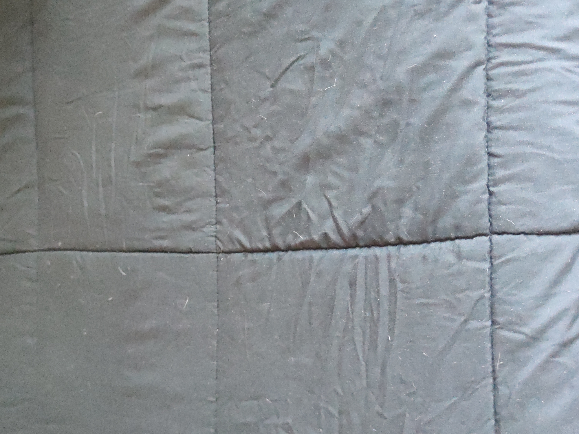The image shows a close-up of a dark gray quilted fabric. The fabric has horizontal and vertical seams forming a grid pattern of squares. The fabric is slightly wrinkled and has some light-colored fibers or lint on its surface. The location is Unknown, Unknown.