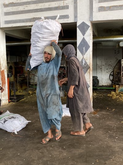 In a manufacturing setting likely in Shurandam, Afghanistan, two men are present. The first man carries a large white bag over his shoulder while wearing a light blue, dirty tunic and sandals. His head is covered with a white cloth. The second man, wearing a dark tunic and sandals, stands next to him, his head covered by a hood. They stand on a concrete floor in a space with industrial machinery visible in the background, as well as bags. The background also features a pillar painted with grey diamond patterns. The light in the room is diffused.